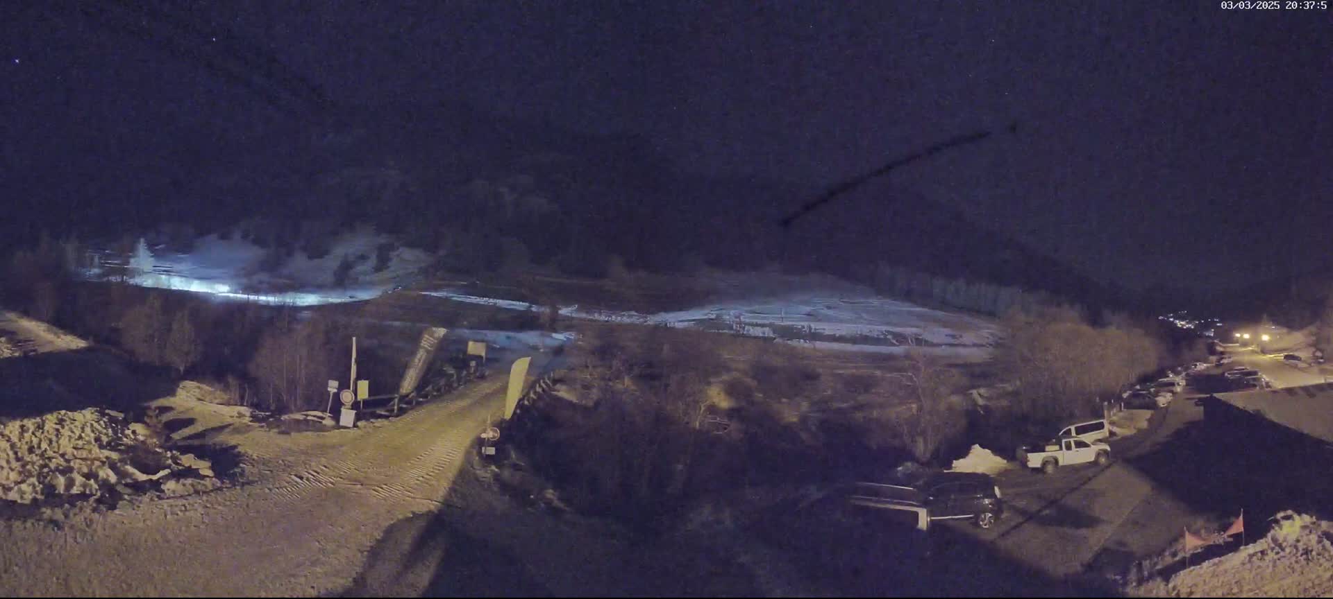 A nighttime, snow-covered ski slope with a parking lot and several vehicles visible under a clear, starry sky.