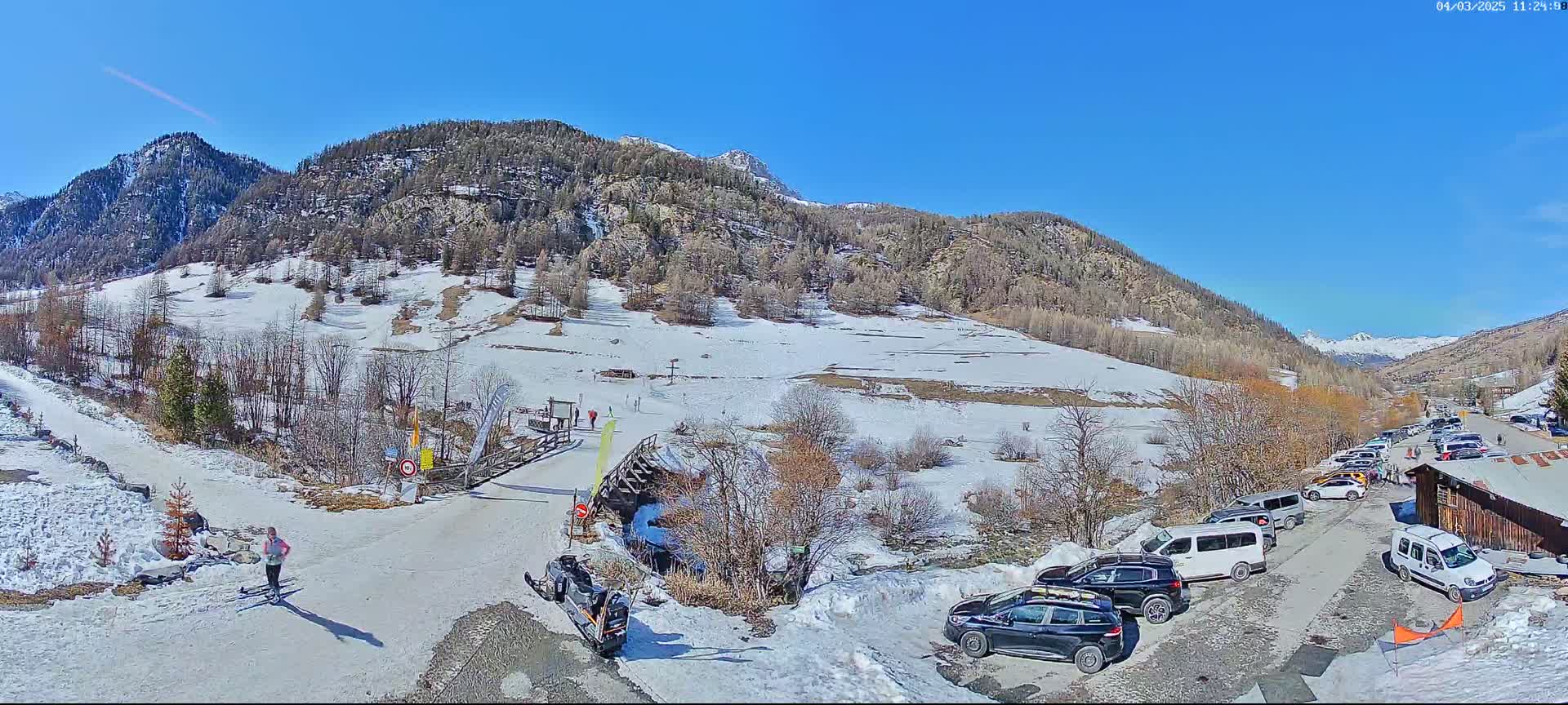 A snow-covered mountain valley scene on a sunny day shows a parking lot with several cars and vans near a small building, a snowmobile, a person cross-country skiing, and a wooden bridge.
