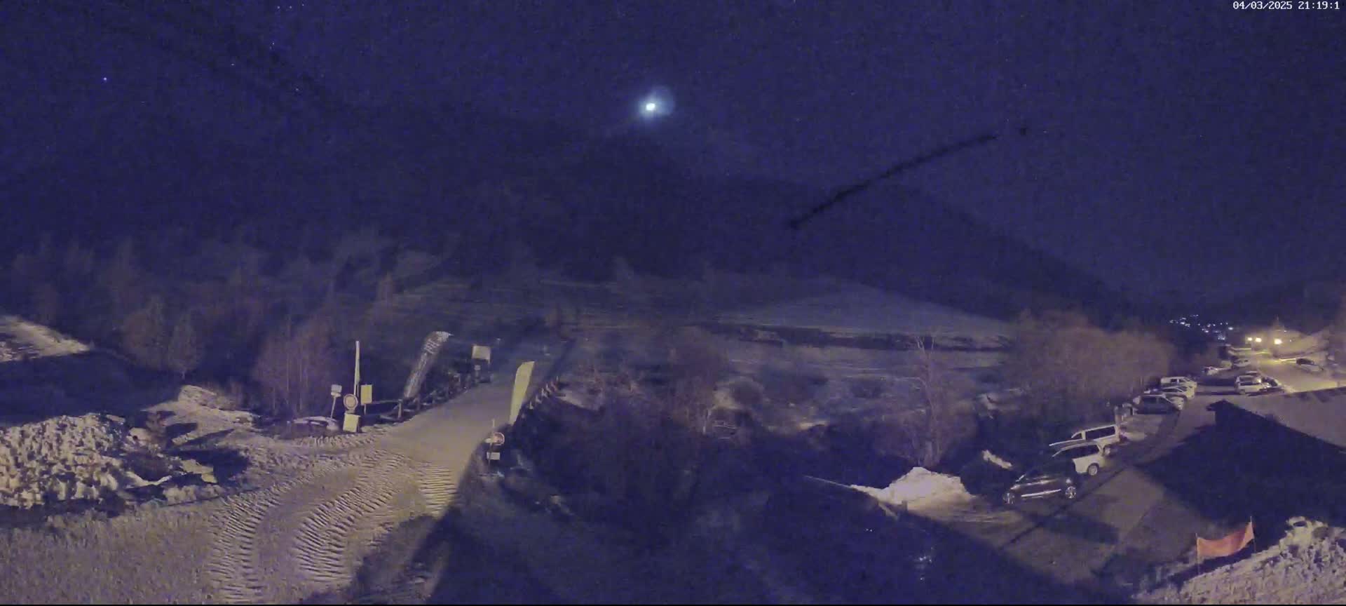A nighttime, snow-covered scene shows a road with parked cars and a bright light in the night sky.