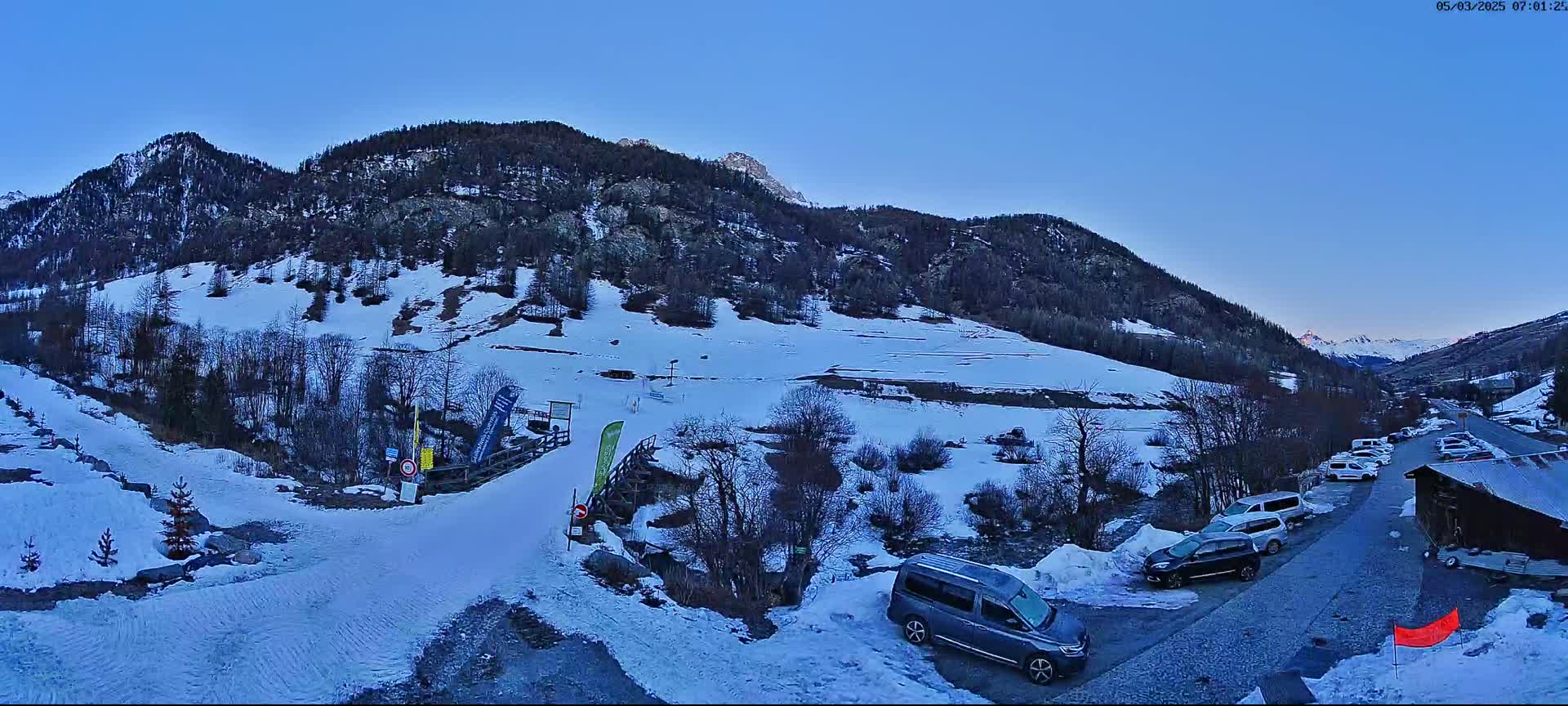 A snow-covered mountainside with several vehicles parked in a lot near a small building under a clear blue sky.