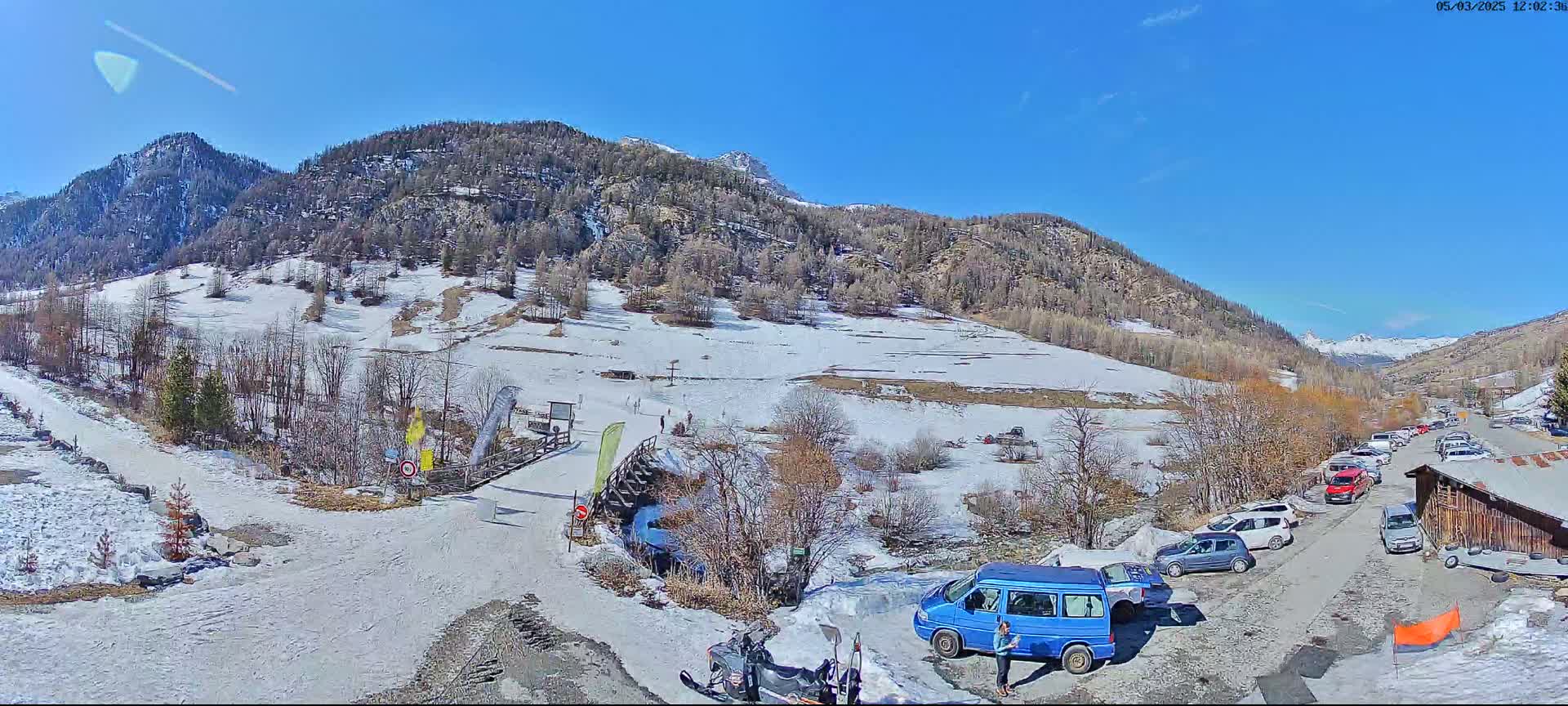 A sunny day in a snow-dusted mountain valley shows a parking lot with several cars and a blue van, near a snow-covered road leading to a mountain pass.