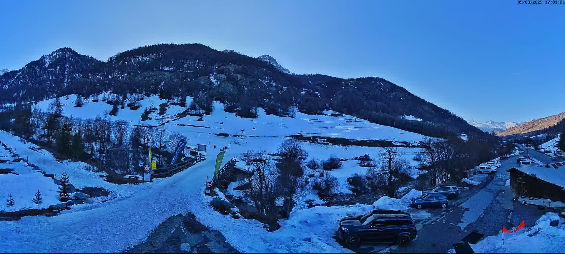 A snow-covered mountain slope with a road leading to a parking lot of cars, under a clear blue sky.