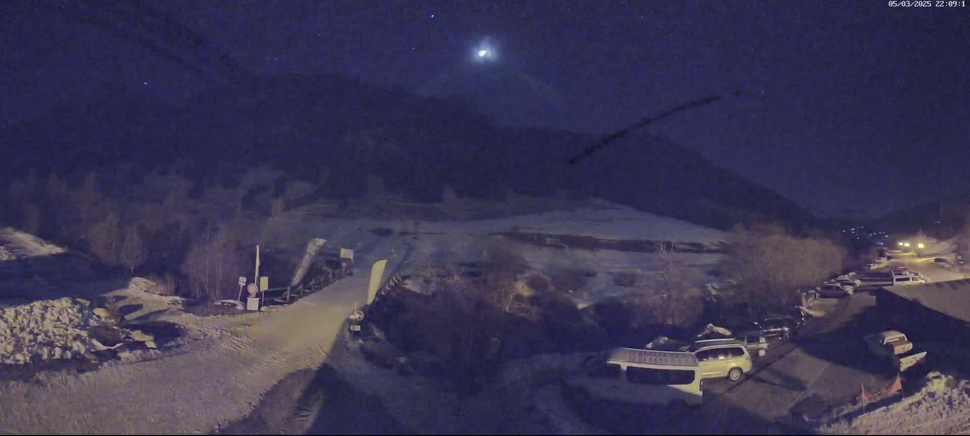 A nighttime, snow-covered landscape shows a parking lot with several vehicles near a mountain under a dark, starry sky with a bright celestial body.