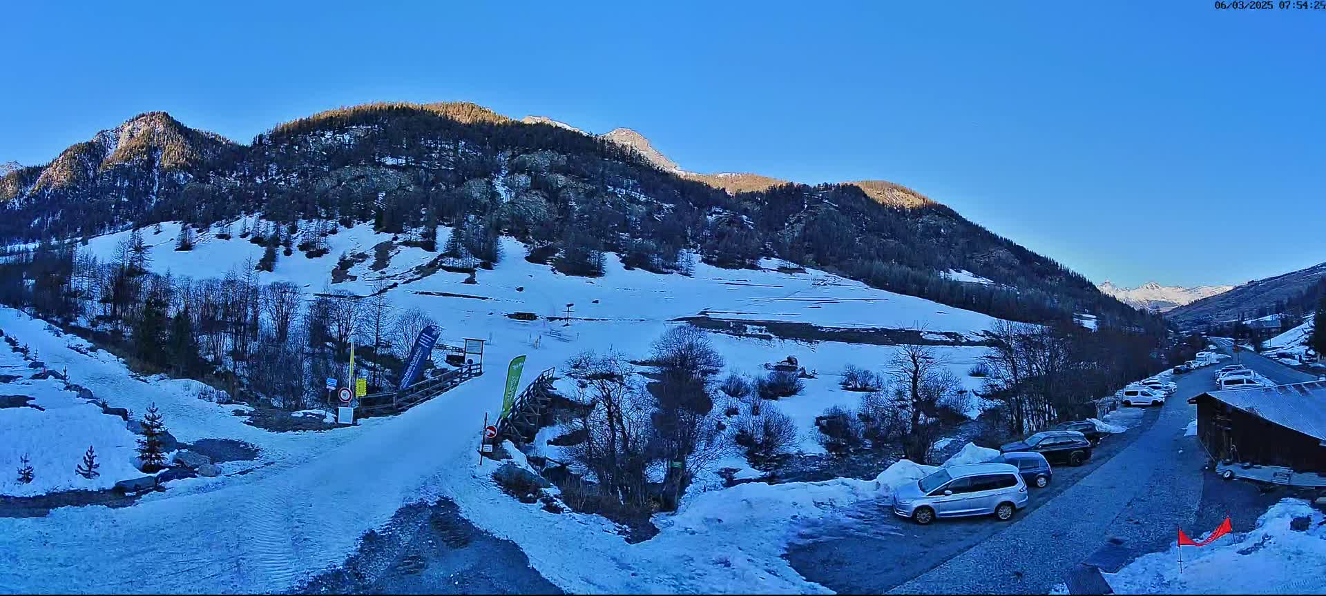 A snow-covered mountainside with a parking lot of cars, a small wooden building and a road leading to a snow-covered area under a clear blue sky.