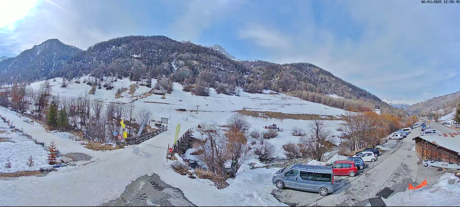 A snow-covered mountain valley scene with a partly sunny sky shows a parking lot with several cars near a wooden building, a snow-covered road leading to a wooden bridge, and a snow-dusted hillside beyond.
