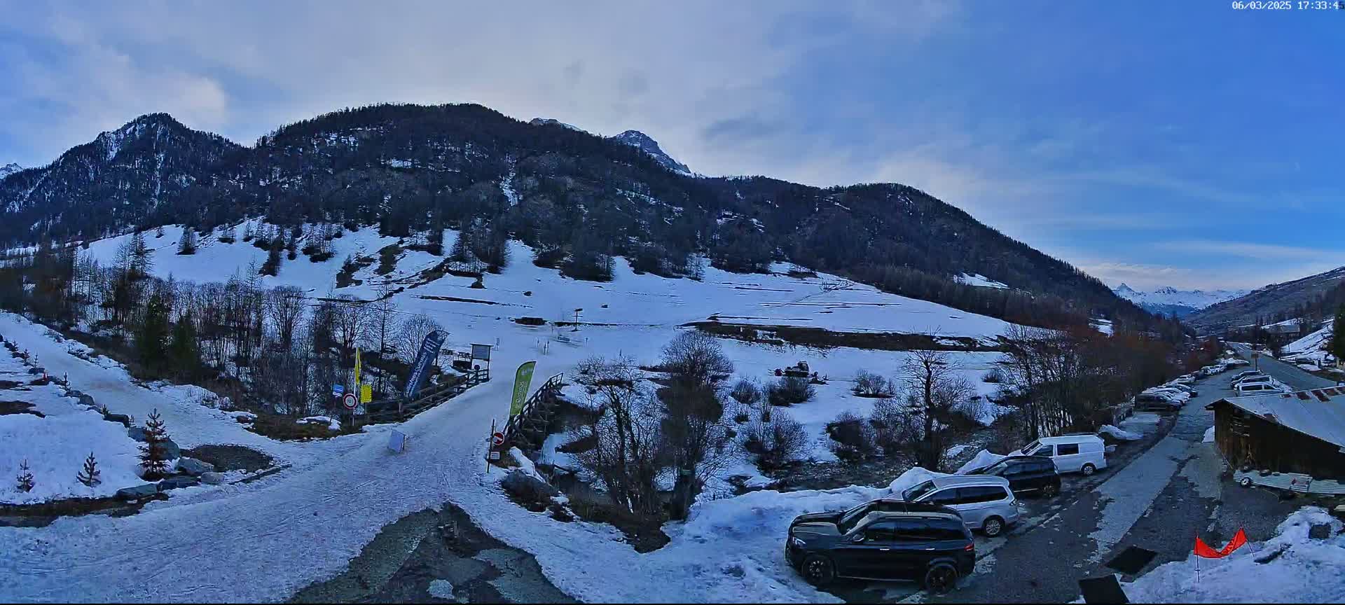 A snow-covered mountain valley with a parking lot containing several cars, under a mostly clear blue sky.
