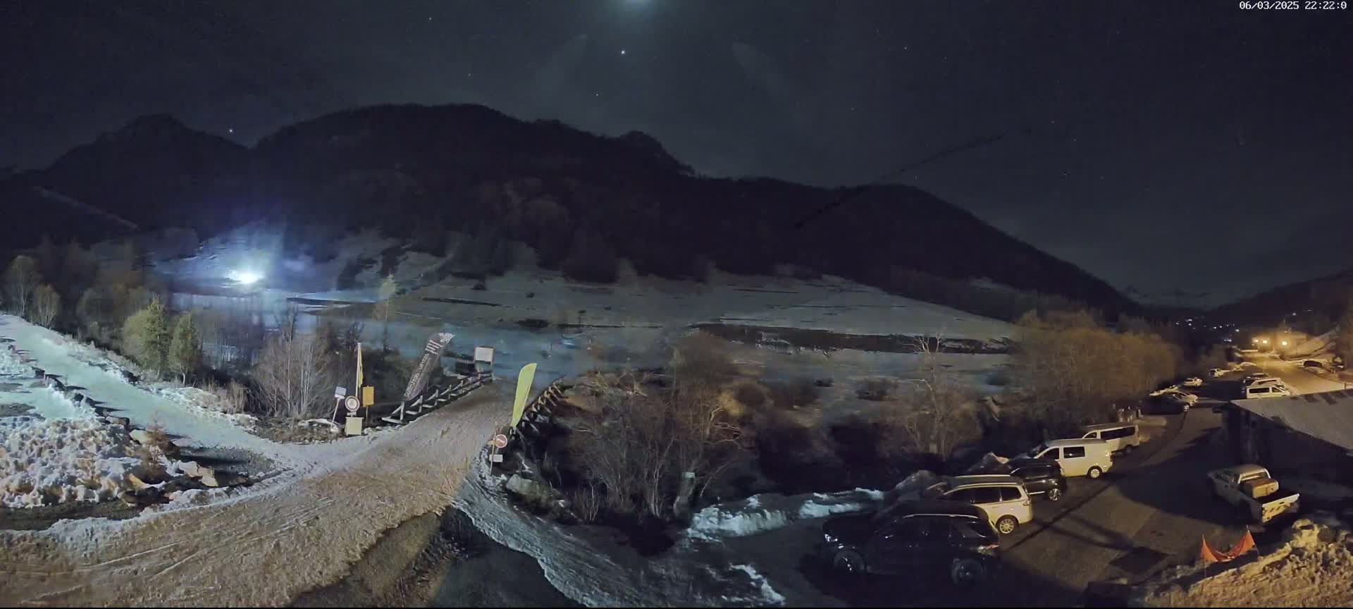 A nighttime view of a snow-dusted mountain valley with several cars parked in a lot near a road leading to a snow-covered area, under a clear, starry sky.