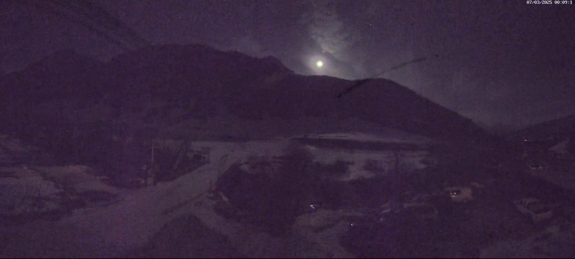 A nighttime view of a mountainous area under a partly cloudy sky with a bright moon.