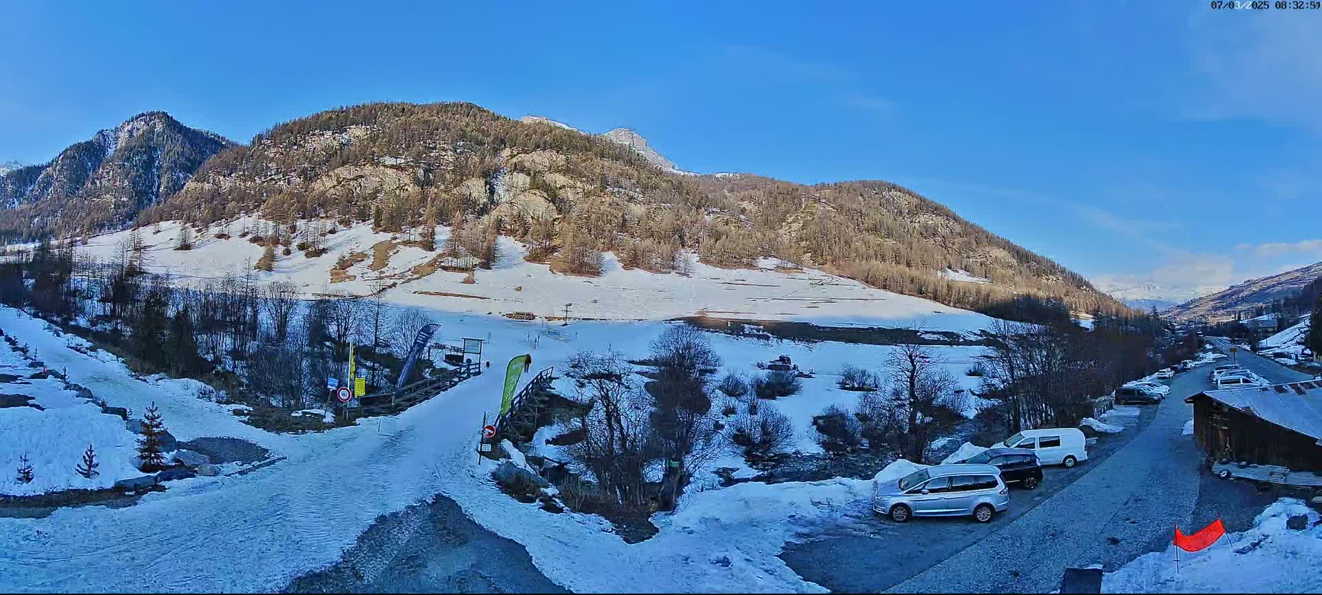 A snow-covered mountain valley scene under a clear blue sky, with several cars parked near a small building and a snow-covered path leading to a bridge.