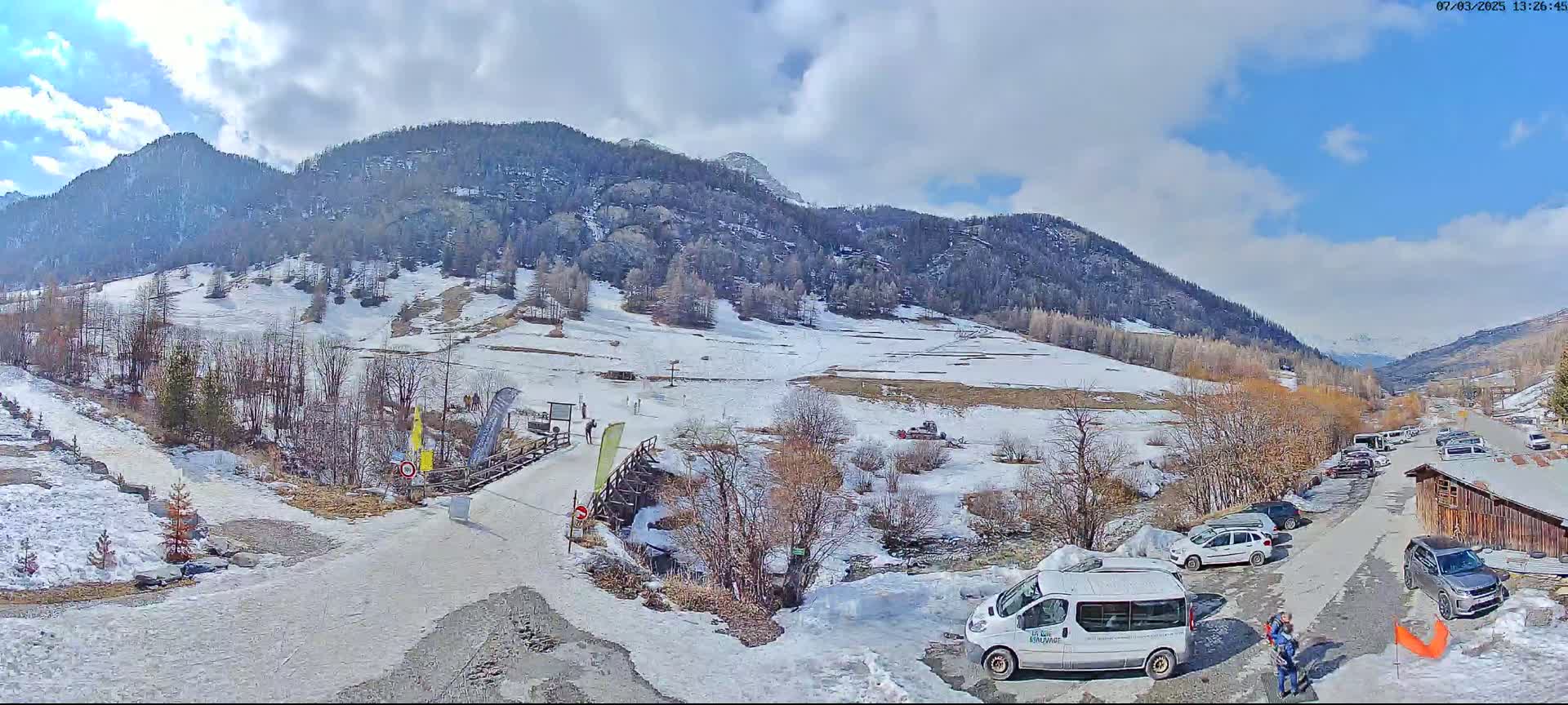 A partly sunny mountain valley scene with snow on the ground and some patches of bare earth, featuring a parking lot with several vehicles, a small wooden building, and a wooden footbridge.