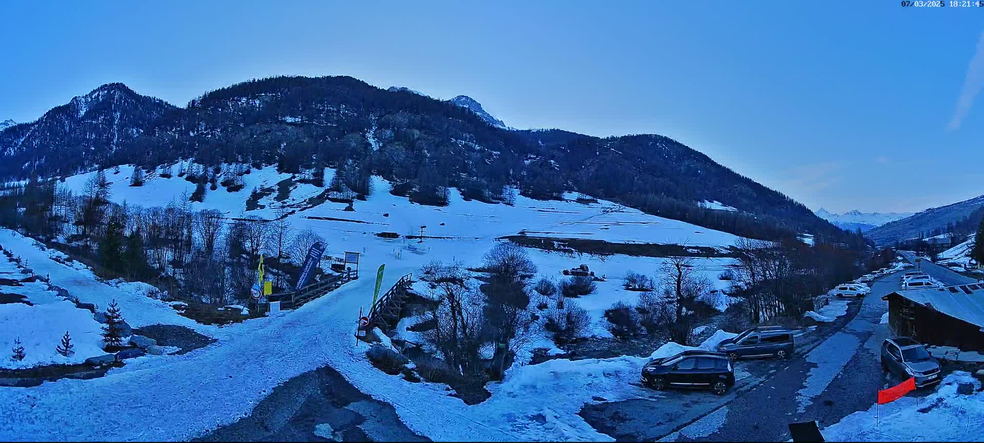 A snow-covered mountain valley at dusk, with cars parked near a wooden building and a snow-covered path leading to a small ski slope.