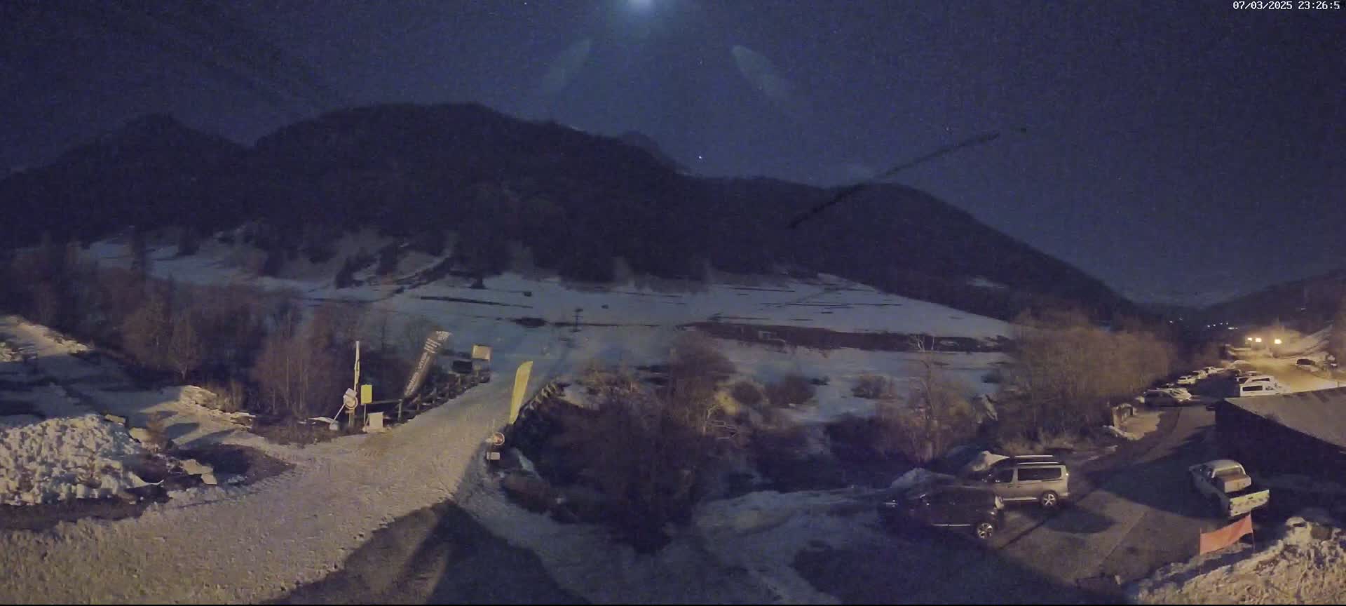 A nighttime view of a snow-covered mountainside with a road leading to a parking lot containing several vehicles, under a clear, moonlit sky.