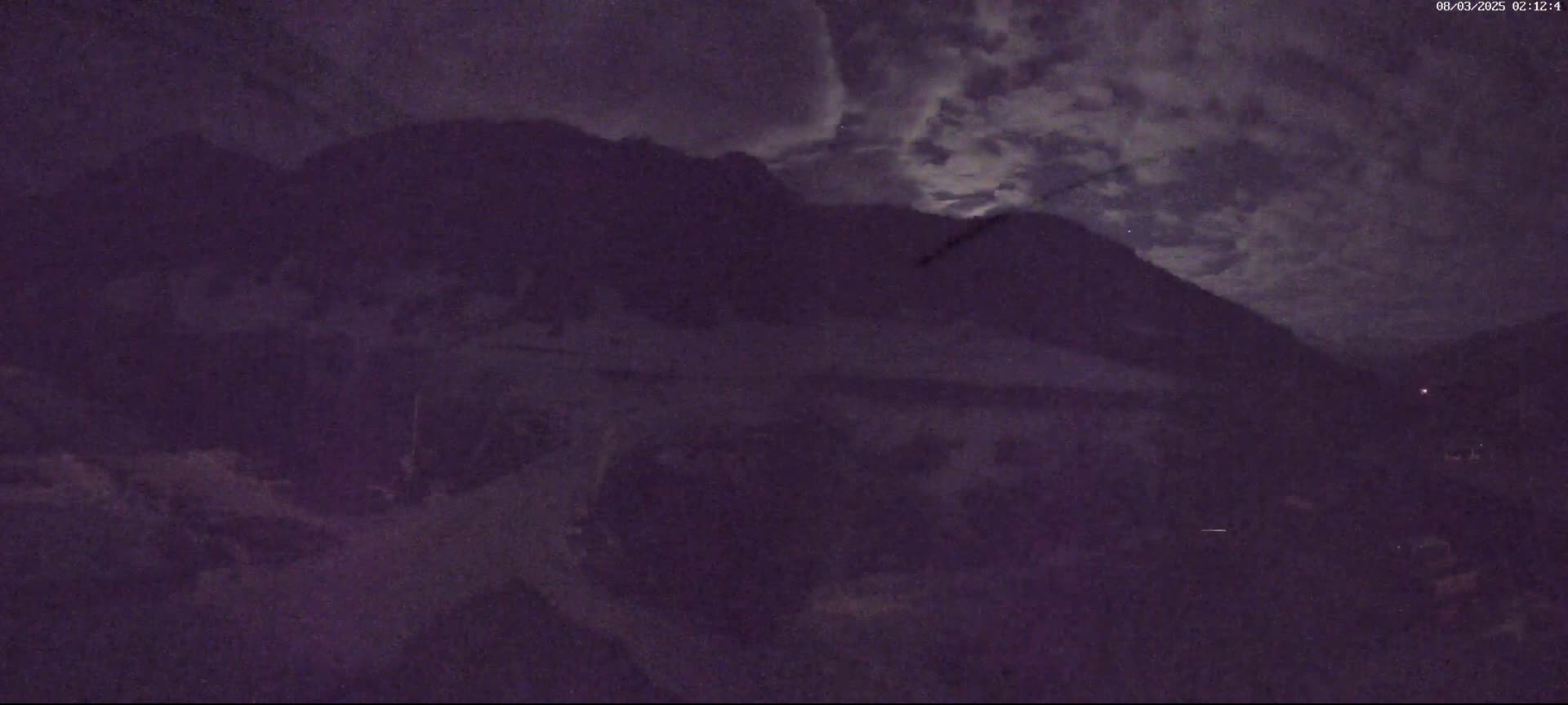 A nighttime view of a mountainous landscape under a cloudy sky with a faint moon visible.