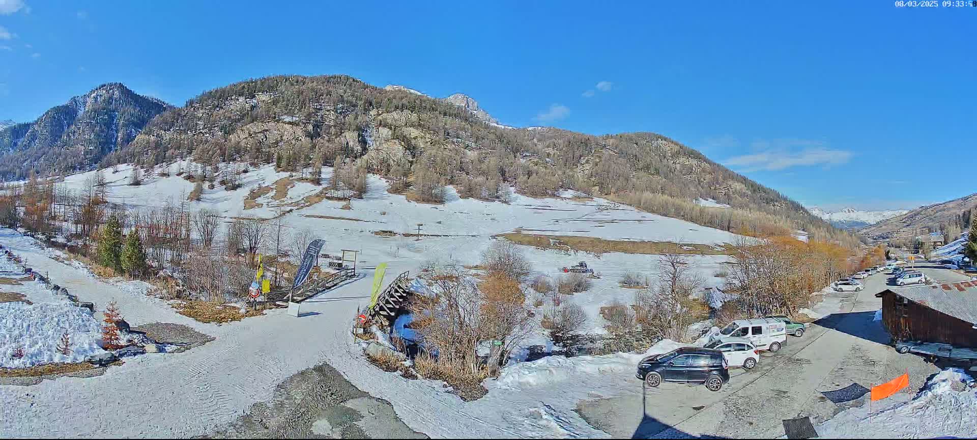 A snow-dusted mountain valley scene under a bright, sunny sky shows a parking lot with several cars near a small building, with a snow-covered ski slope and trees leading up to the mountains in the background.