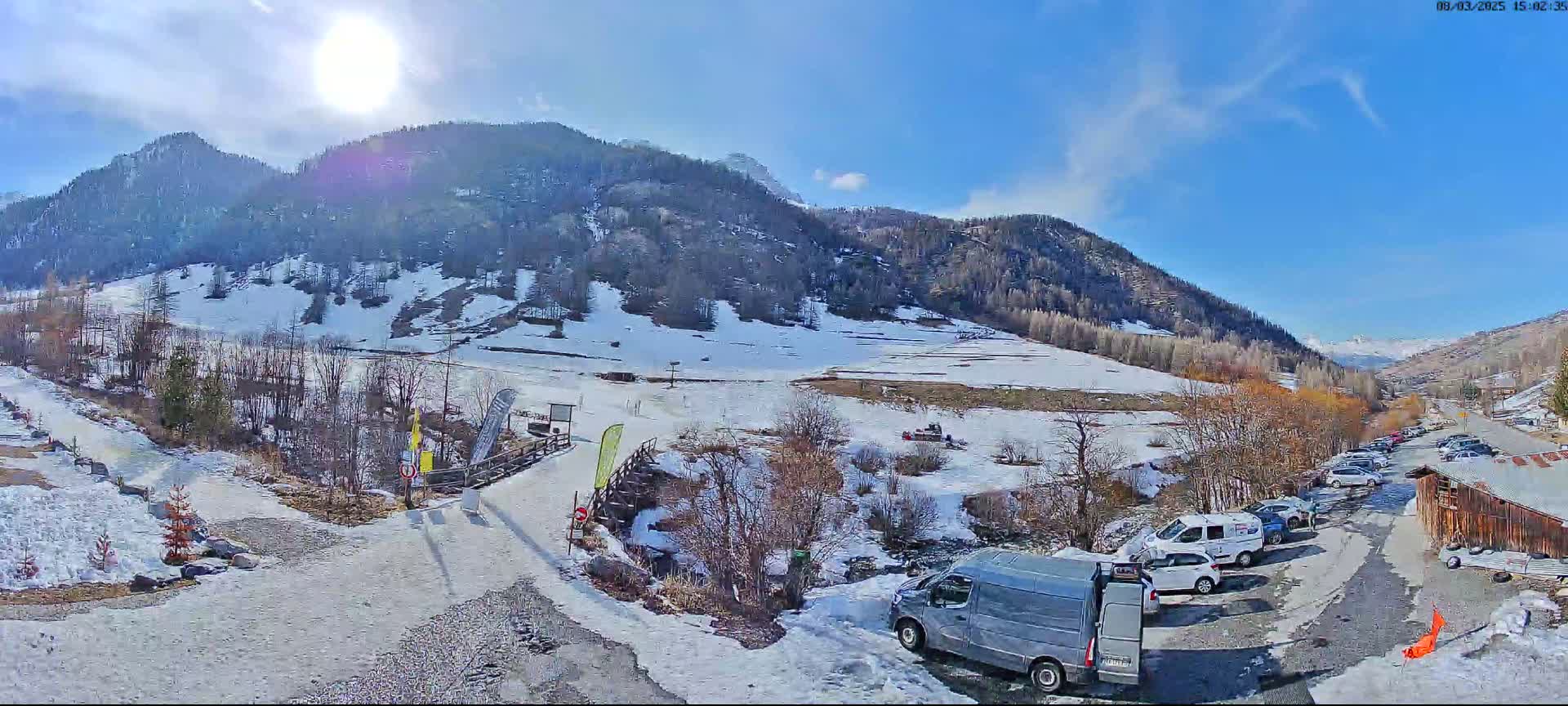A sunny, snow-covered mountain valley scene shows a parking lot with several vehicles near a small wooden building, with a snow-dusted path leading to a wooden bridge and a ski area in the background.