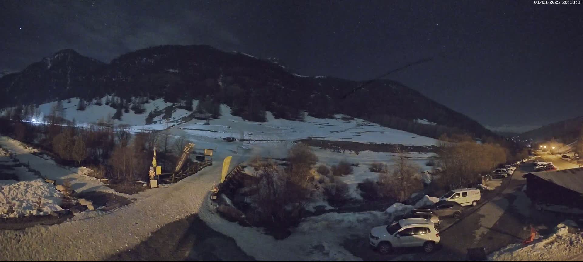 A snow-covered mountainside at night, with a parking lot and several cars visible near the bottom, under a clear, starry sky.