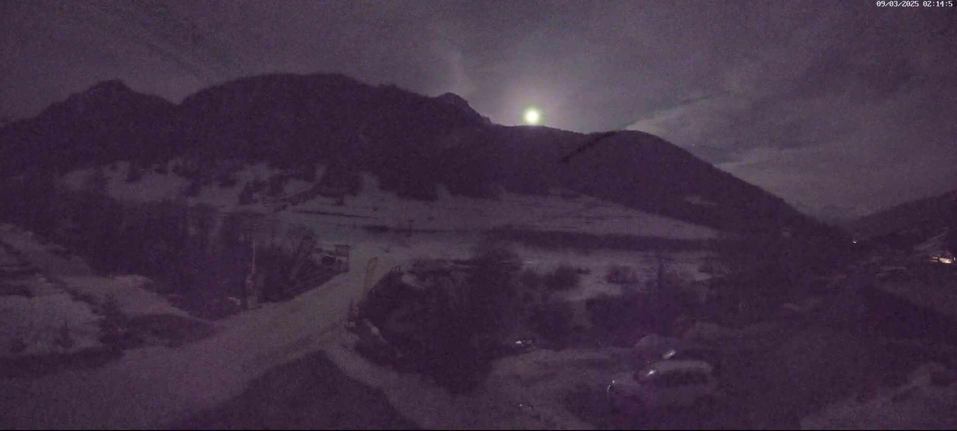 A nighttime view of snow-covered mountains under a bright moon and cloudy sky, with some vehicles and machinery visible in the foreground.