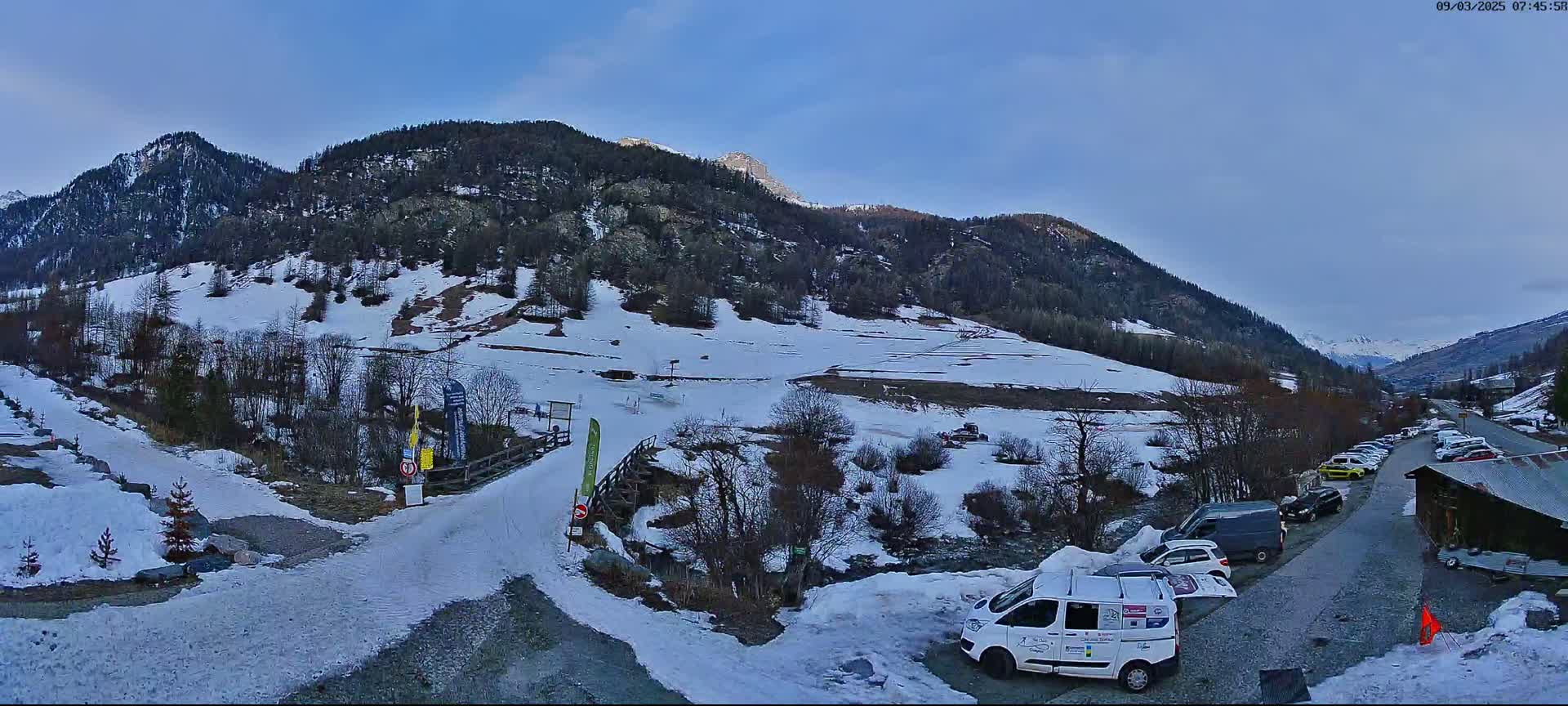 A snow-covered mountain valley with a parking lot full of cars near a road leading to a small ski area, under a clear, light blue sky.