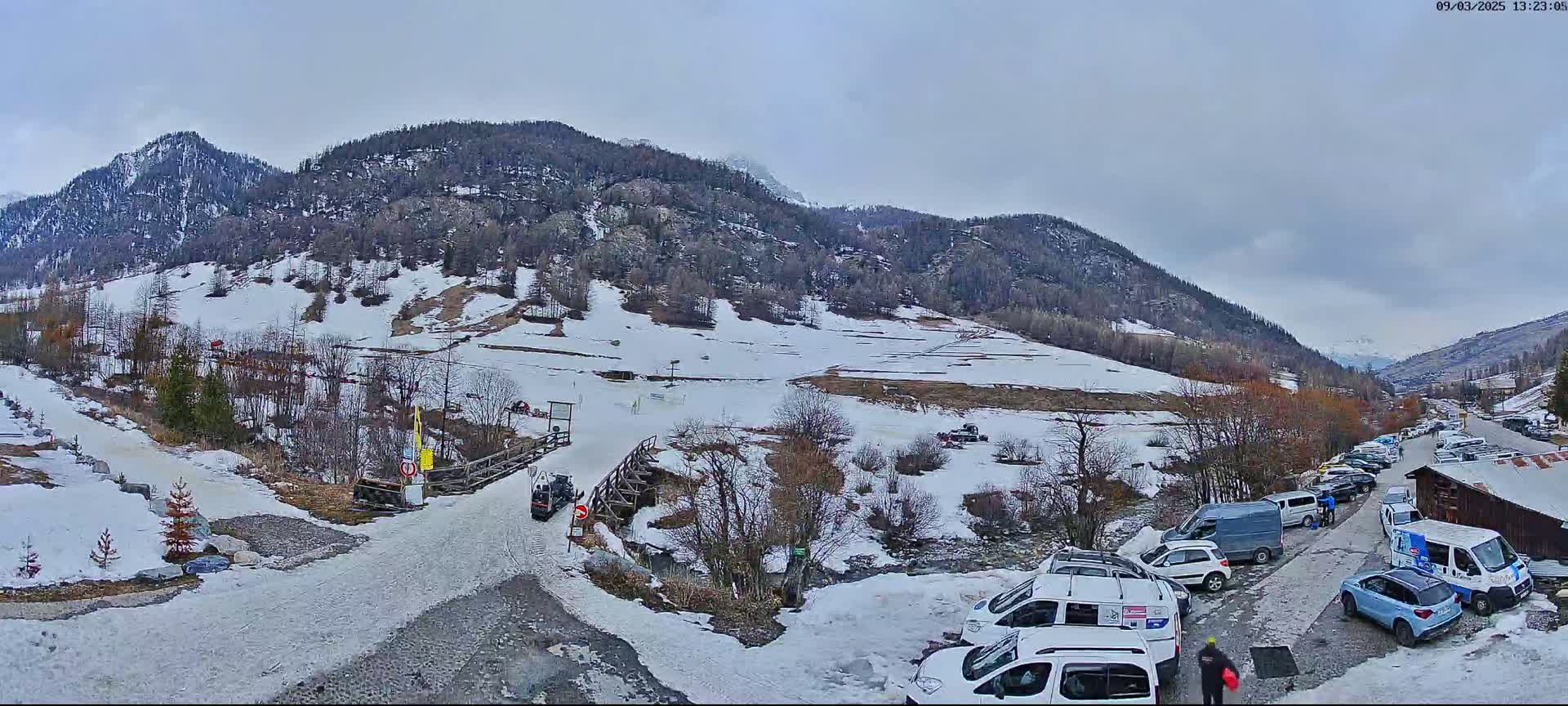 A snow-covered mountain valley with a parking lot full of vehicles, under an overcast sky.