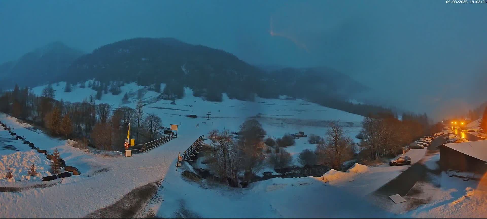 A snow-covered valley at dusk, with a small wooden bridge, a few cars parked near a building, and mountains in the background under a dark blue sky.
