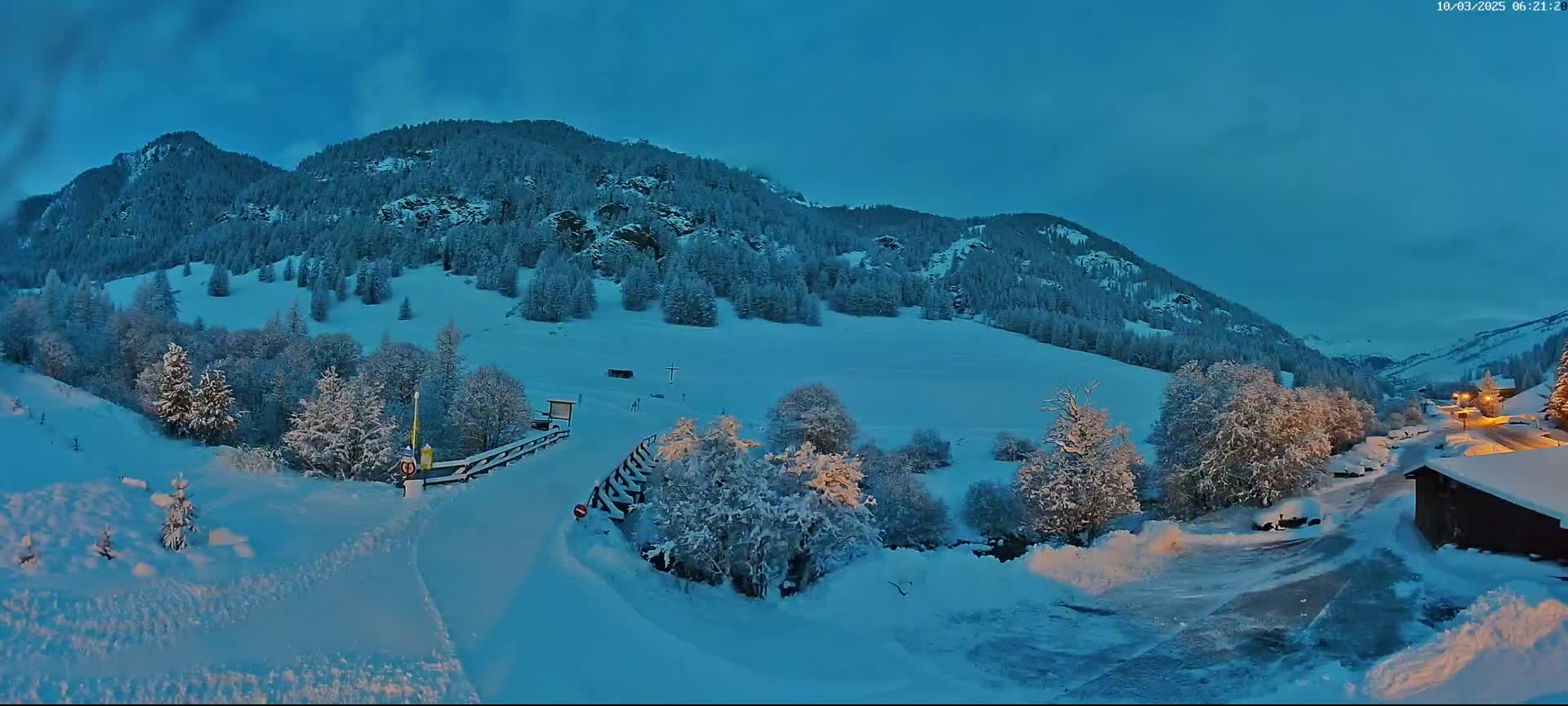 A snow-covered mountain valley at twilight, with a road leading to a small village nestled among snow-laden trees.
