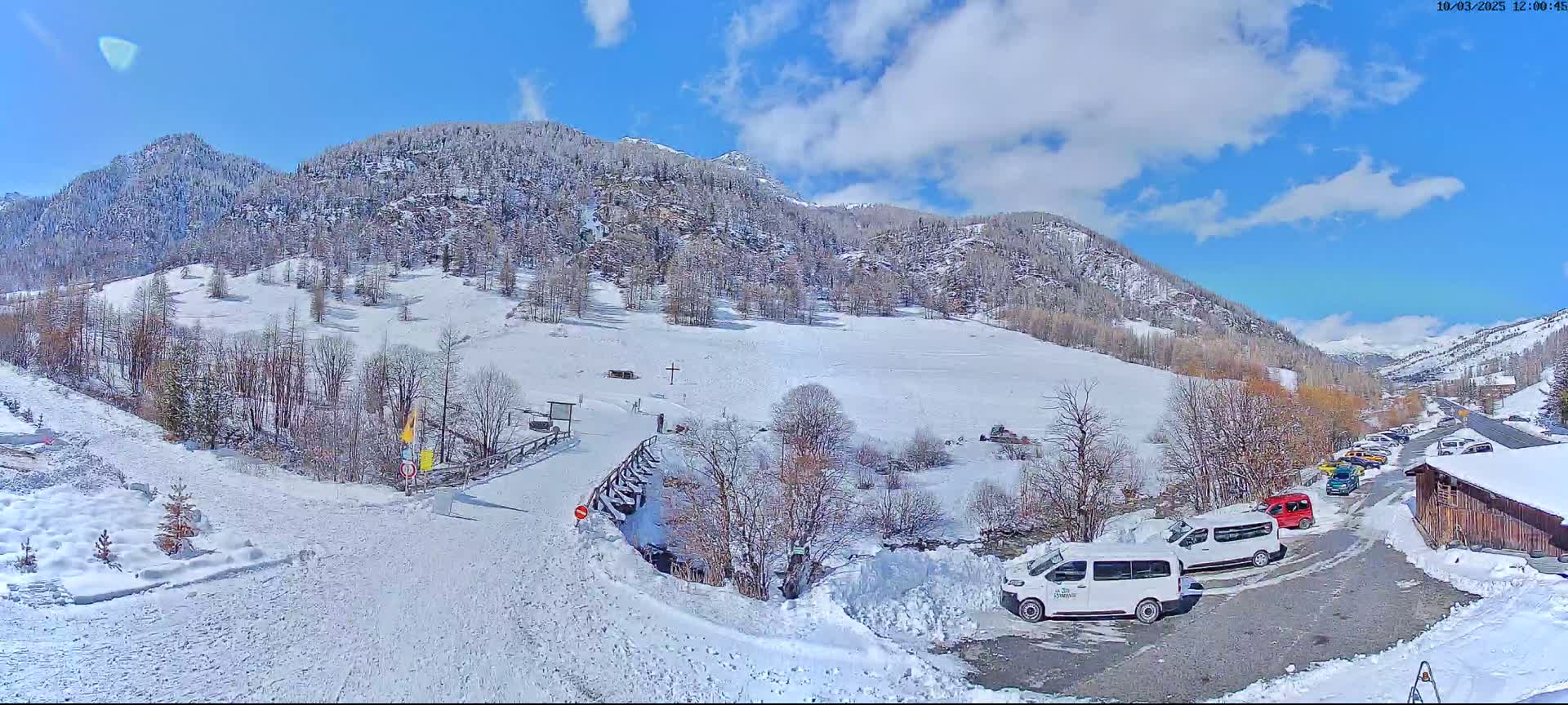 A snow-covered mountain valley with a few vehicles parked near a wooden building under a partly sunny sky.