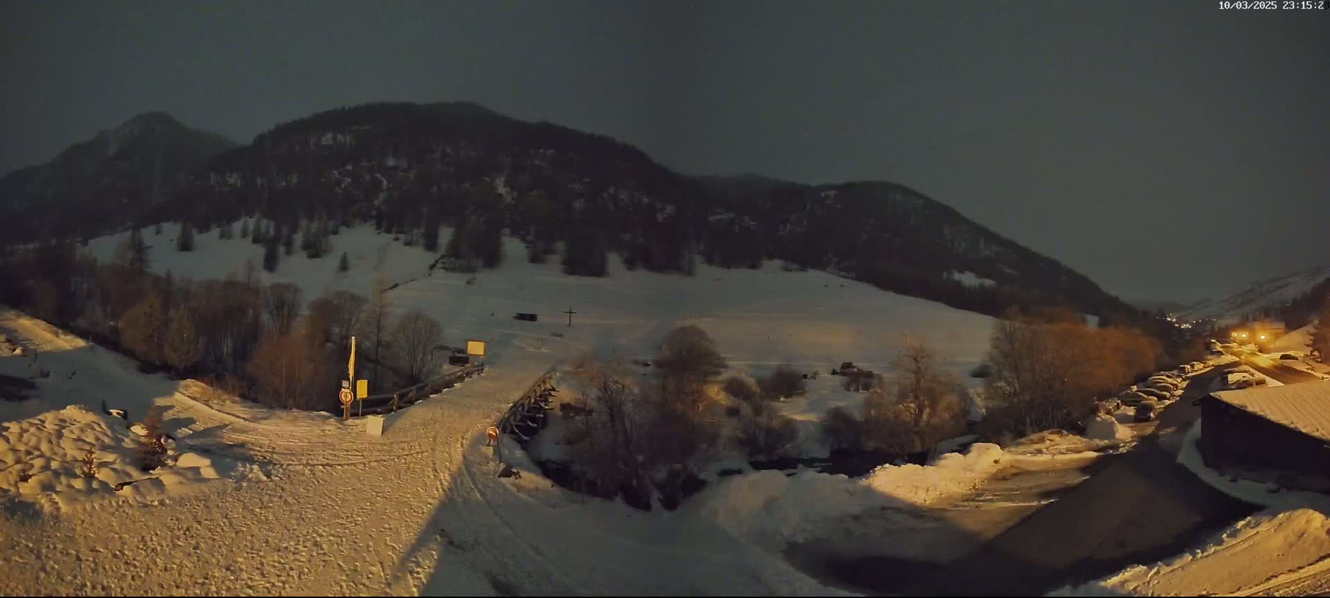 A snow-covered landscape at night, illuminated by artificial lights, shows a valley nestled between mountains under a dark sky.