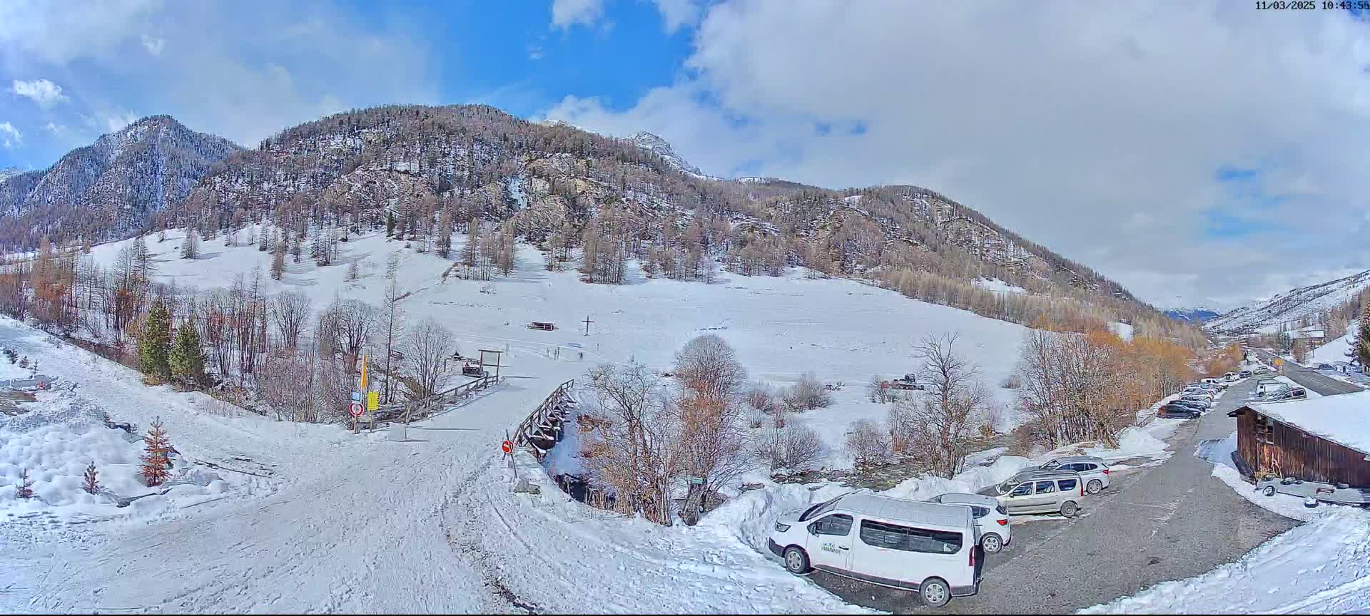 A snow-covered valley with sparsely wooded hillsides, a wooden bridge, several parked cars, and a partly sunny sky.