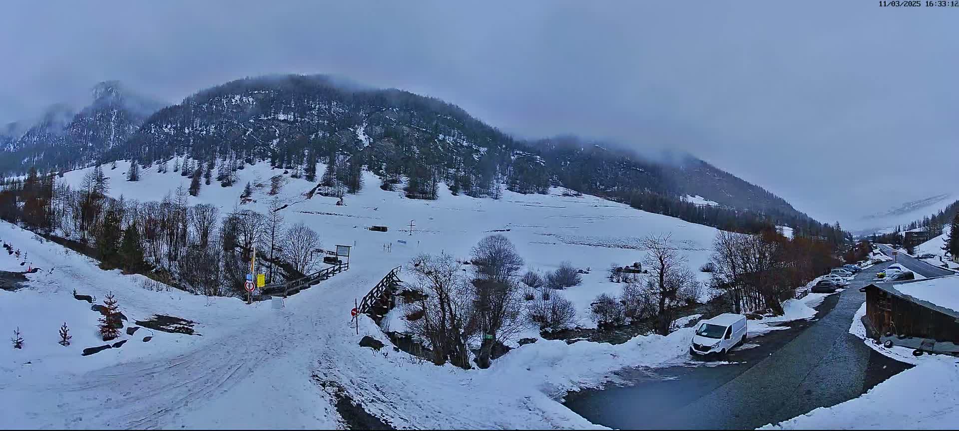 A snowy mountain valley scene on an overcast day, with a road leading to a small parking area and a few buildings nestled among snow-covered trees.