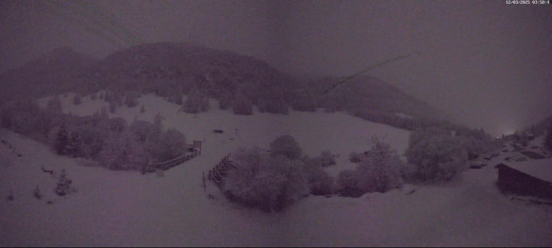 A snow-covered landscape at night, with snow falling, shows a hilly area with trees and a few buildings in the distance.