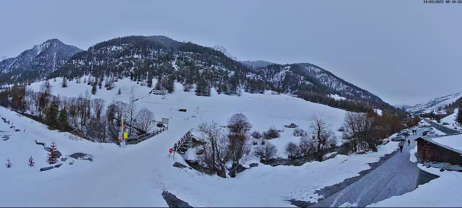 A snow-covered valley nestled between snow-dusted mountains under a cloudy sky, with a small road and a few cars visible.