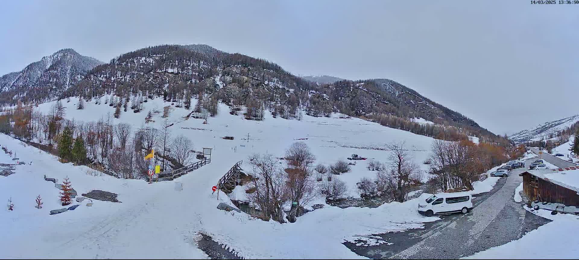 A snow-covered mountain valley scene on an overcast day shows a road leading to a small parking area near a wooden building, with several vehicles visible.