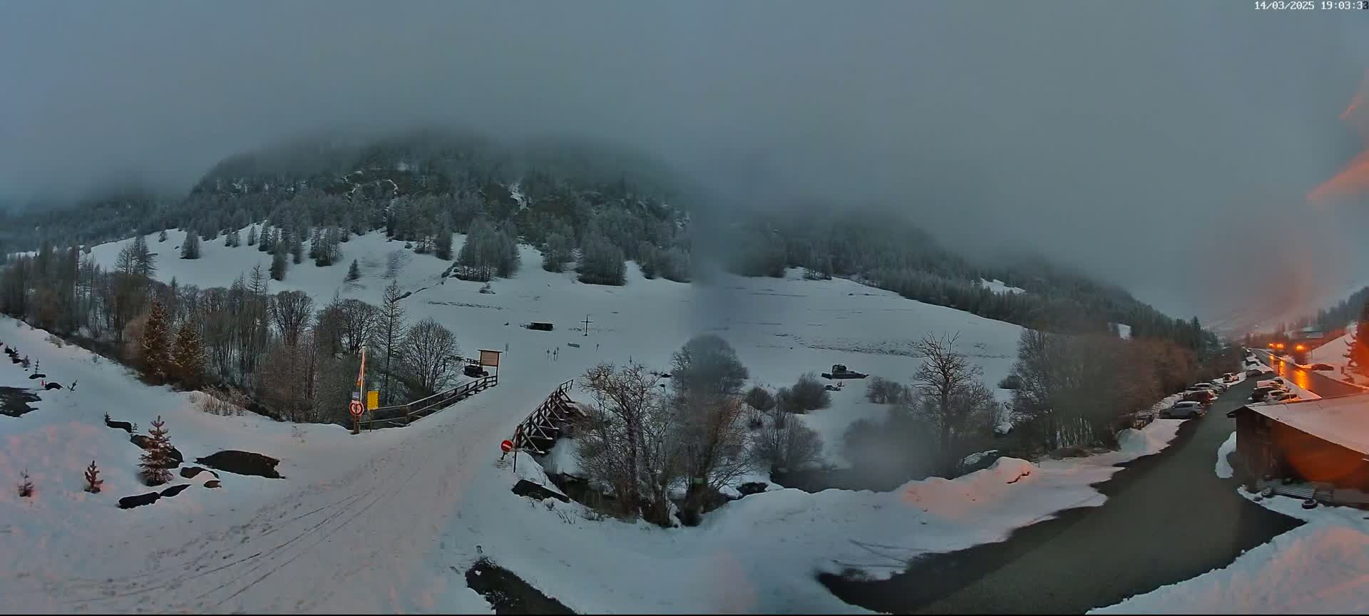 A snow-covered mountain valley is shrouded in fog under a dim, overcast sky, with a road and a small parking lot visible in the foreground.