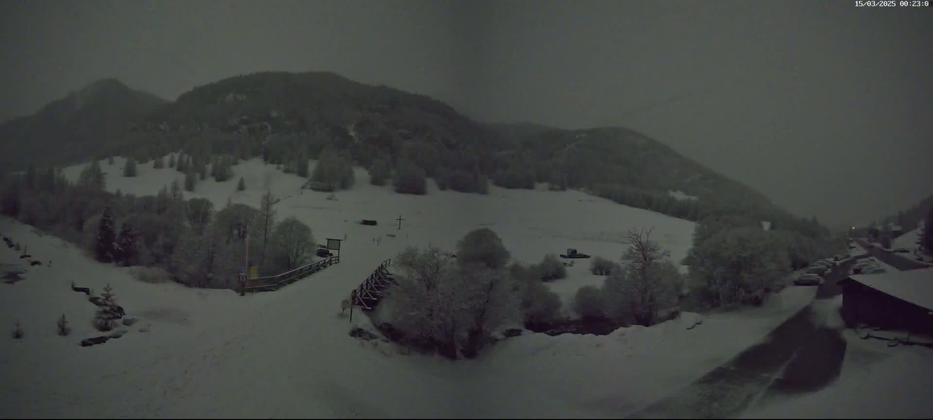 A snow-covered landscape at night, showing a valley with snow-dusted trees and mountains under a dark, overcast sky.