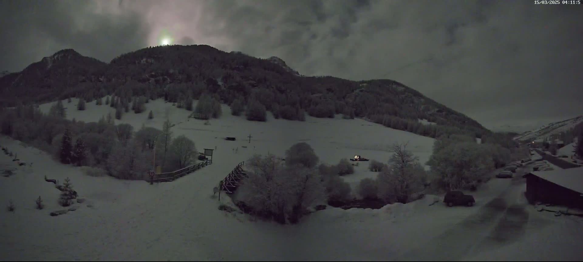 A snow-covered landscape at night, under a dark, cloudy sky, shows a valley with dark trees and mountains in the background.