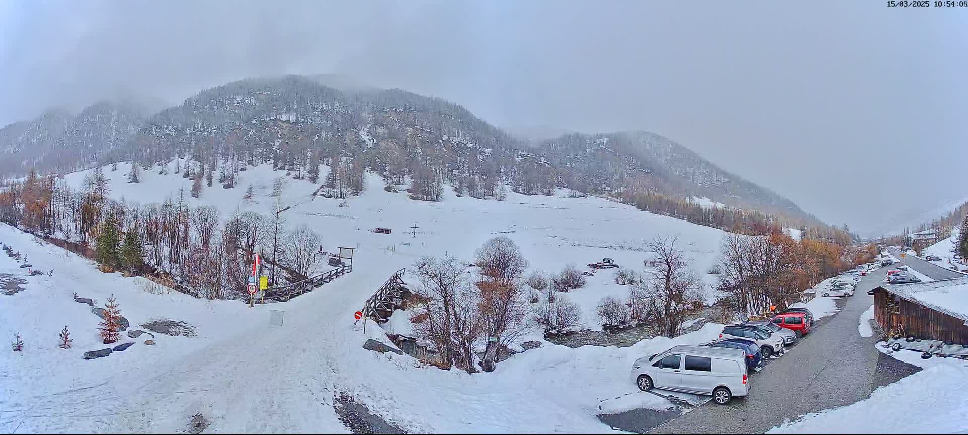 A snowy mountain valley scene shows a parking lot with several cars and a van near a wooden building, with a snow-covered road leading to a wooden bridge and a snow-covered hillside beyond.