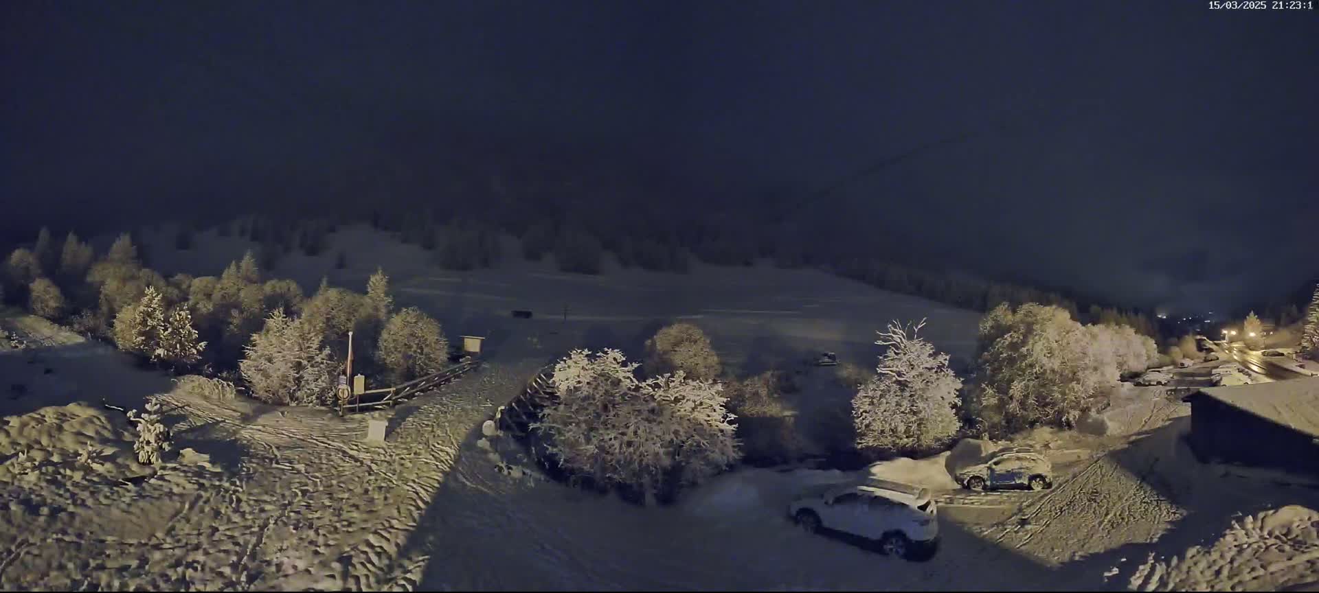 A snow-covered landscape at night, with several vehicles parked near snow-laden trees and a small building.