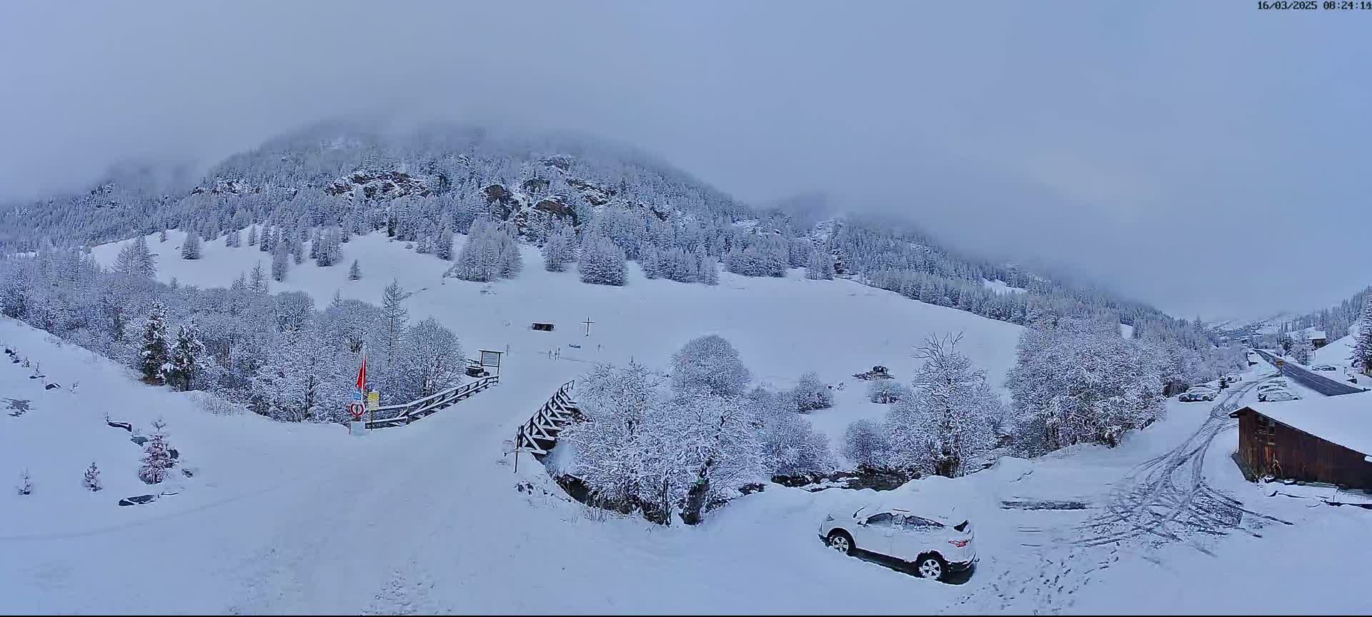 A snow-covered mountain valley scene shows a road leading to a small building, with snow-laden trees and a white SUV parked nearby, under an overcast sky.