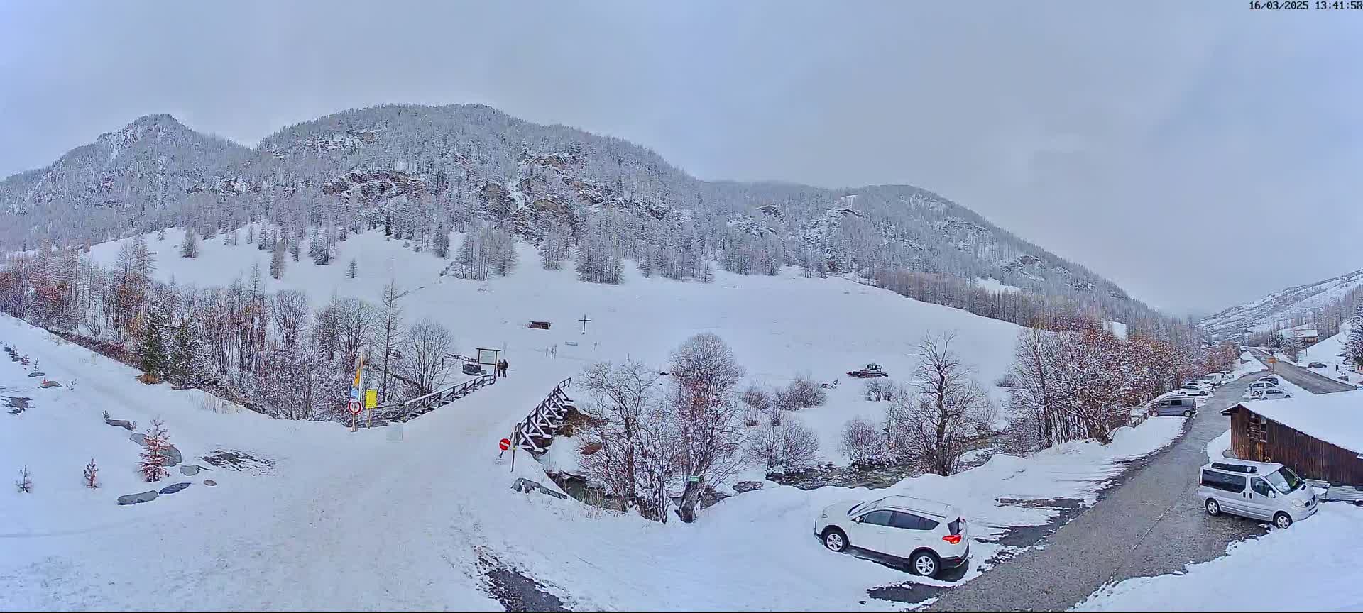A snow-covered mountain valley scene shows a roadway with parked vehicles, a small bridge, and snow-laden trees under an overcast sky.