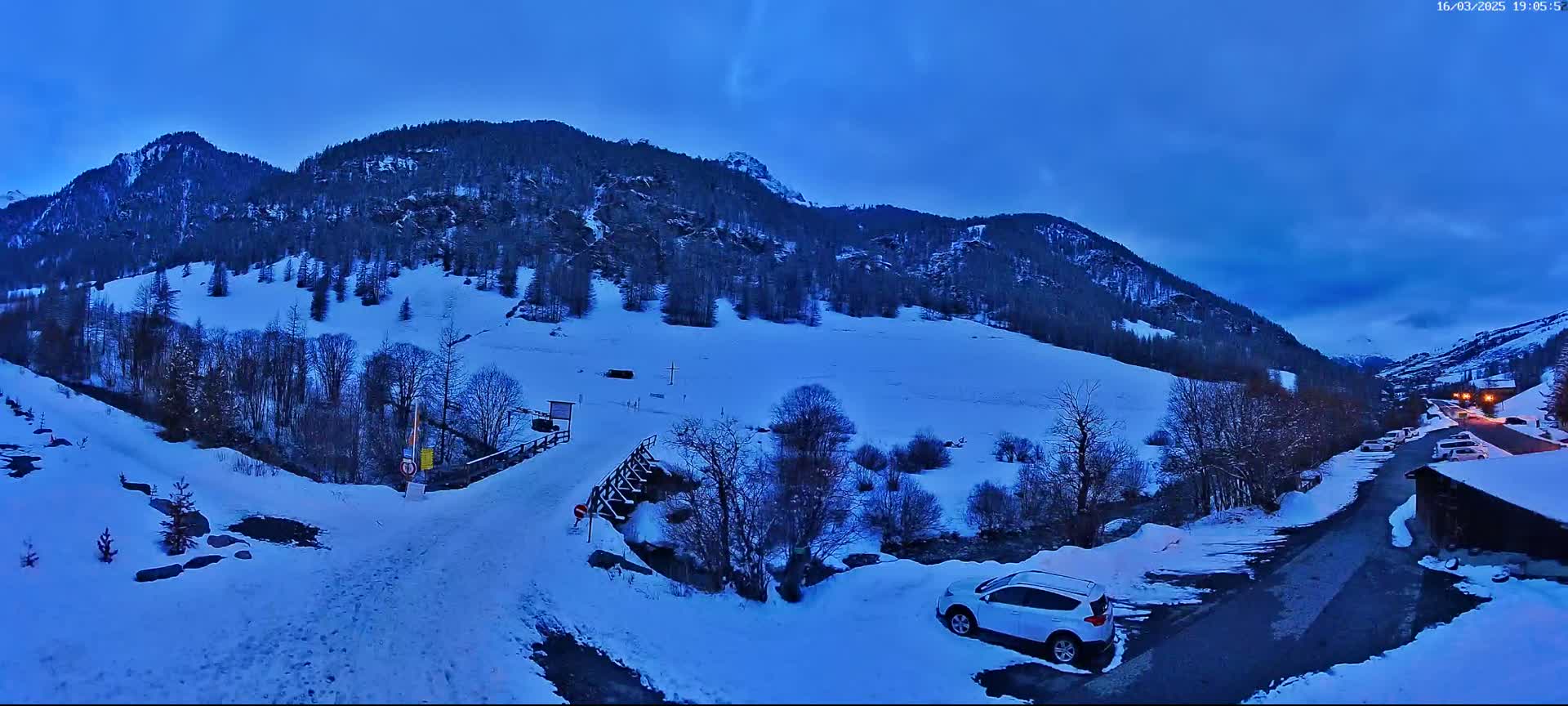 A snow-covered mountain valley at twilight, with a white SUV parked near a road and a small bridge.