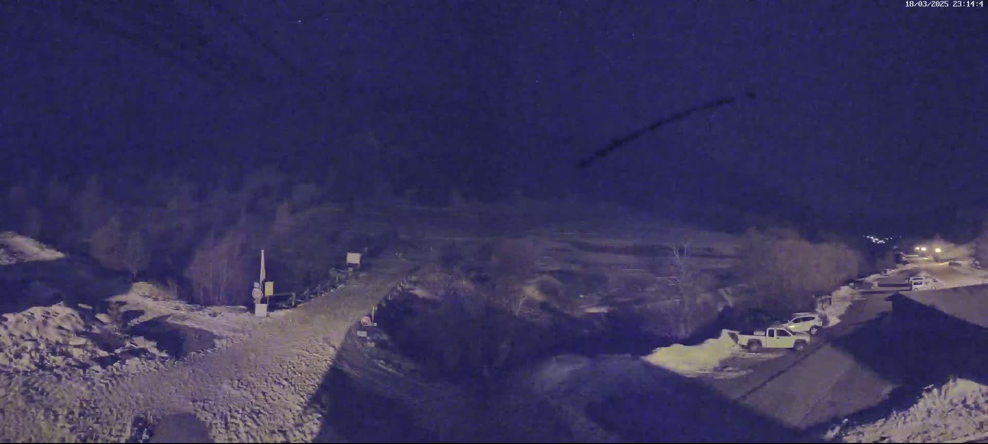 A nighttime, snow-covered landscape shows a road winding through a sparsely vegetated area with several parked vehicles visible under a dark sky.