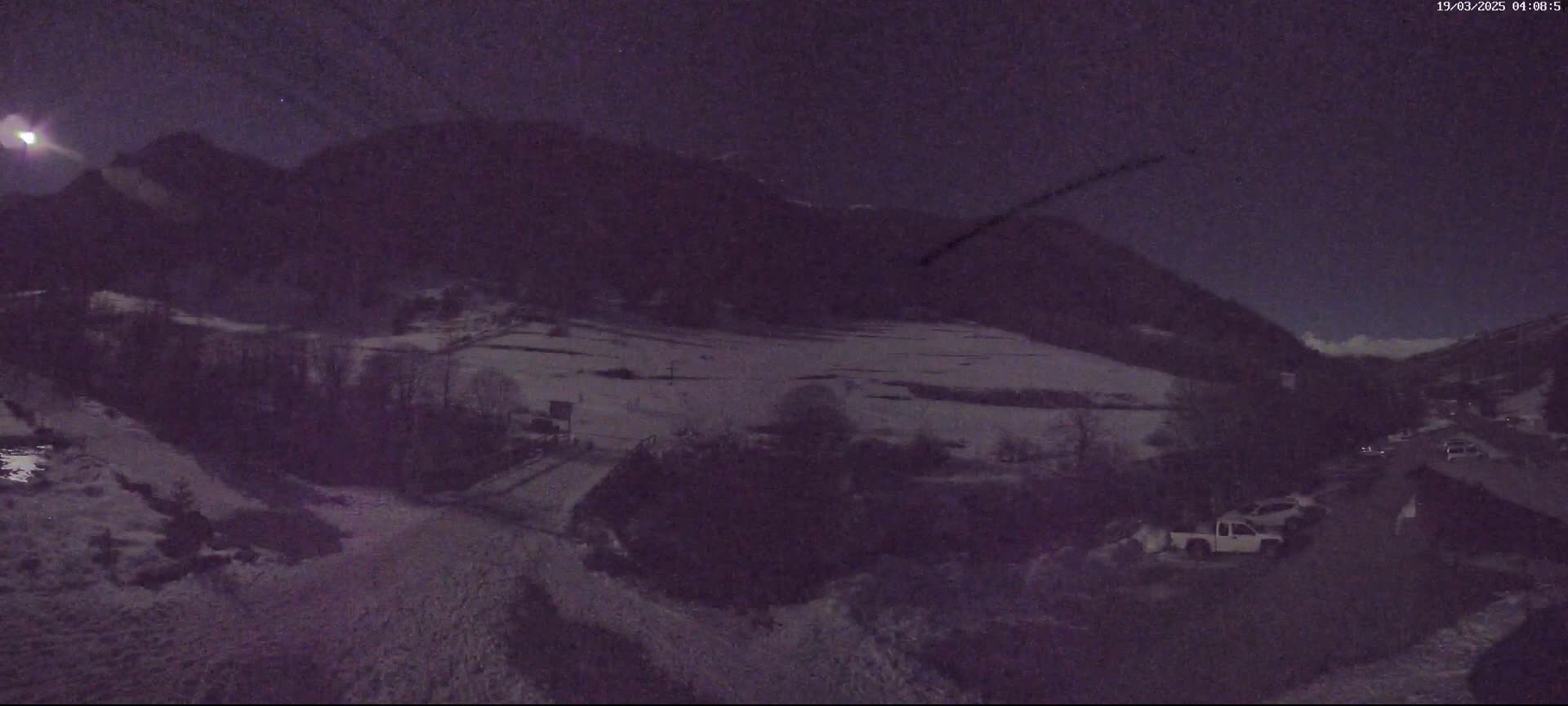 A nighttime view of a snow-covered valley nestled between dark mountains under a partly cloudy moonlit sky.