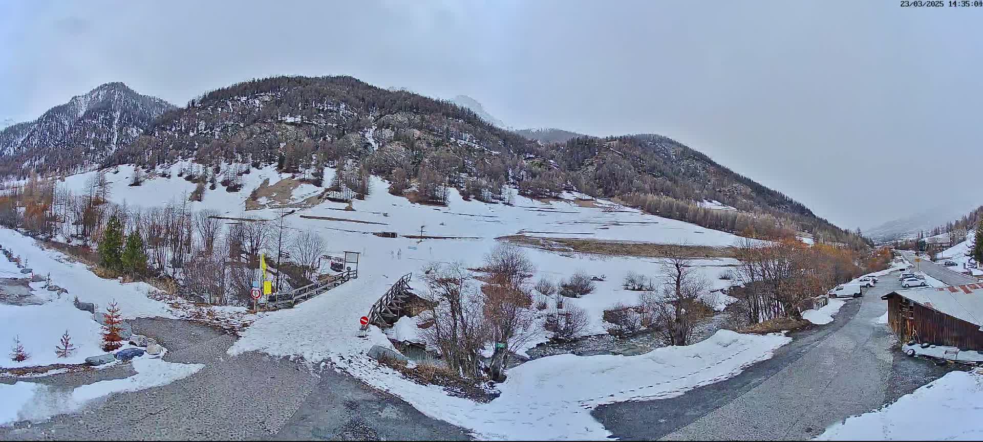 A snow-covered valley with a small wooden bridge, a few cars parked nearby, and mountains in the background under an overcast sky.