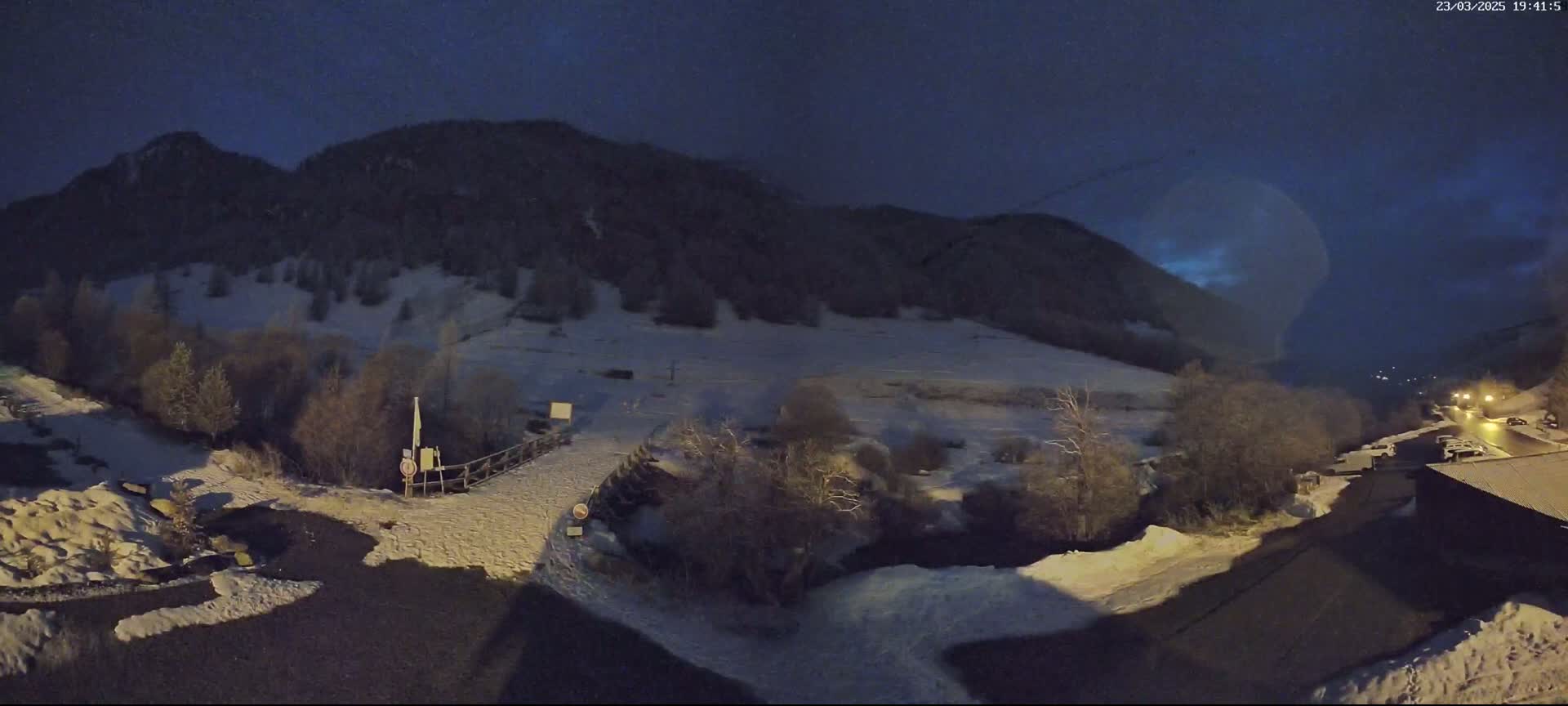 A snow-covered landscape at night, featuring a mountain in the background and a road leading to a building in the foreground under a dark, mostly clear sky.