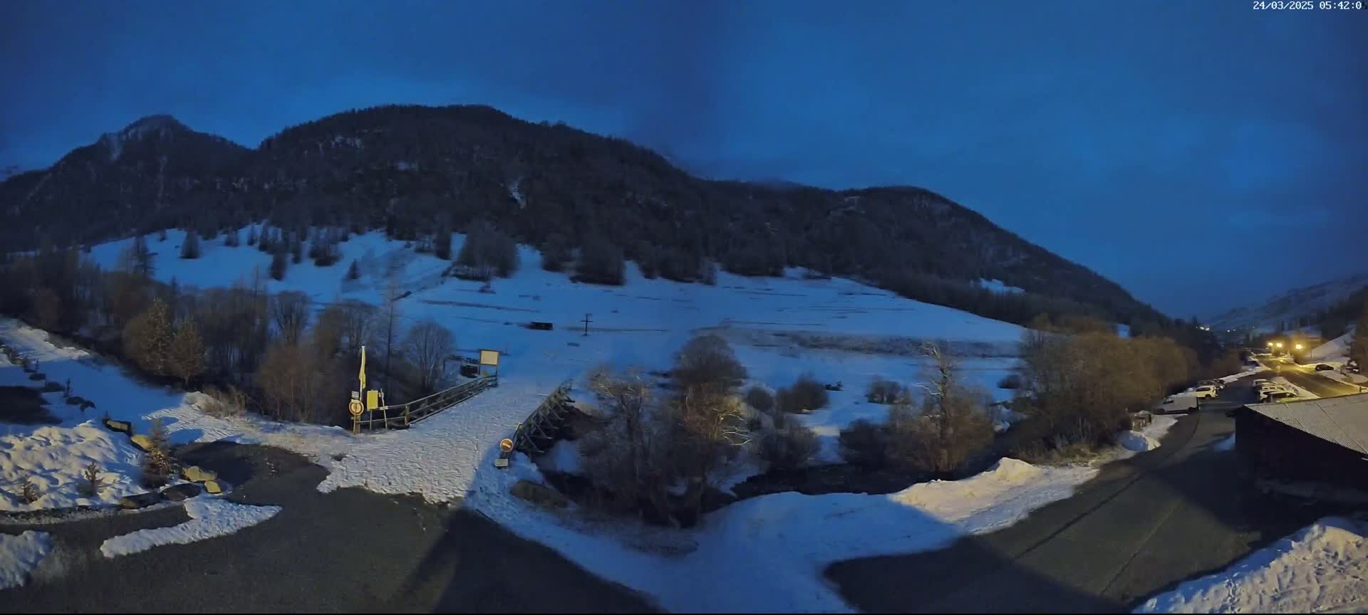 A snow-covered valley at twilight, with a small wooden bridge over a stream, is nestled between dark, snow-dusted mountains.