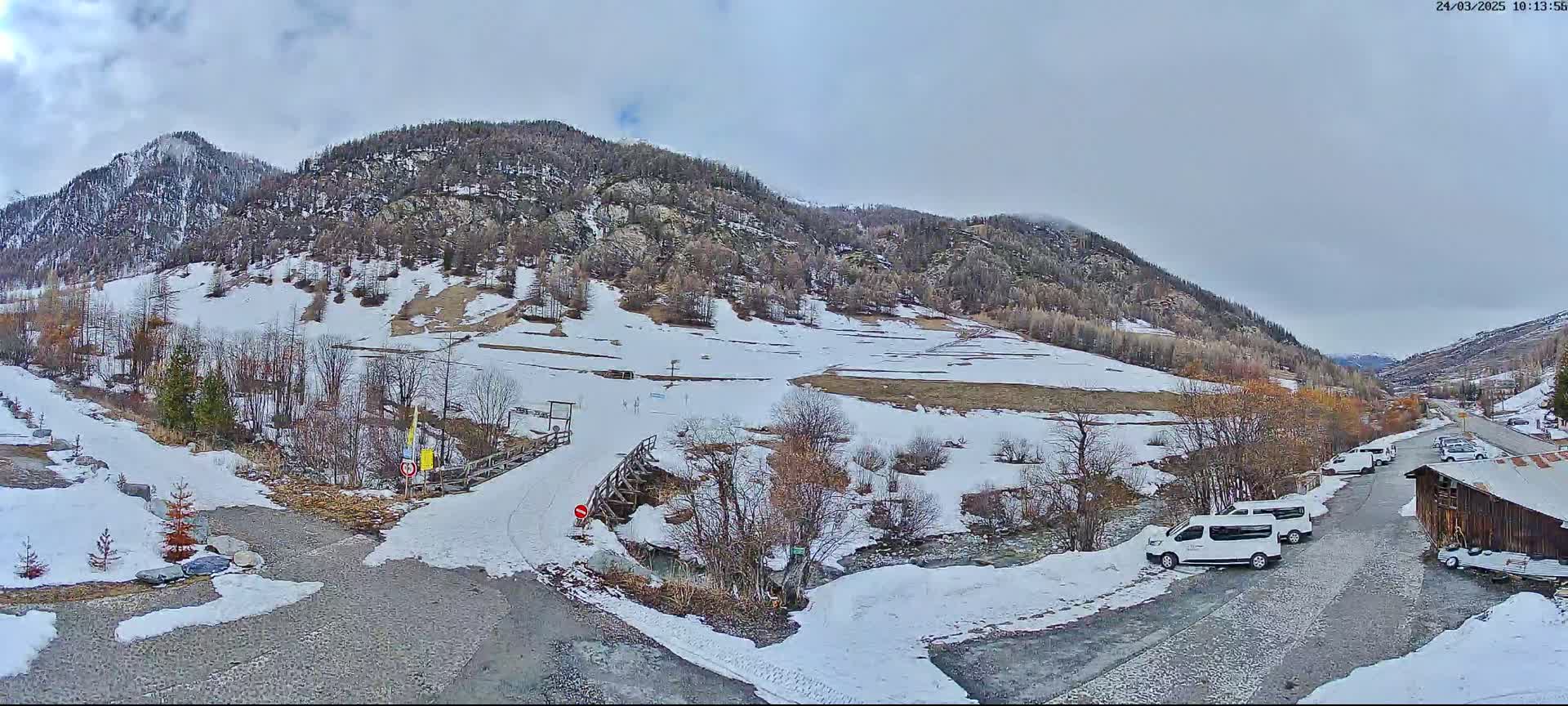 A snow-dusted mountain valley under a cloudy sky features a small parking area with several vehicles near a wooden building, with a wooden bridge crossing a small stream in the foreground.