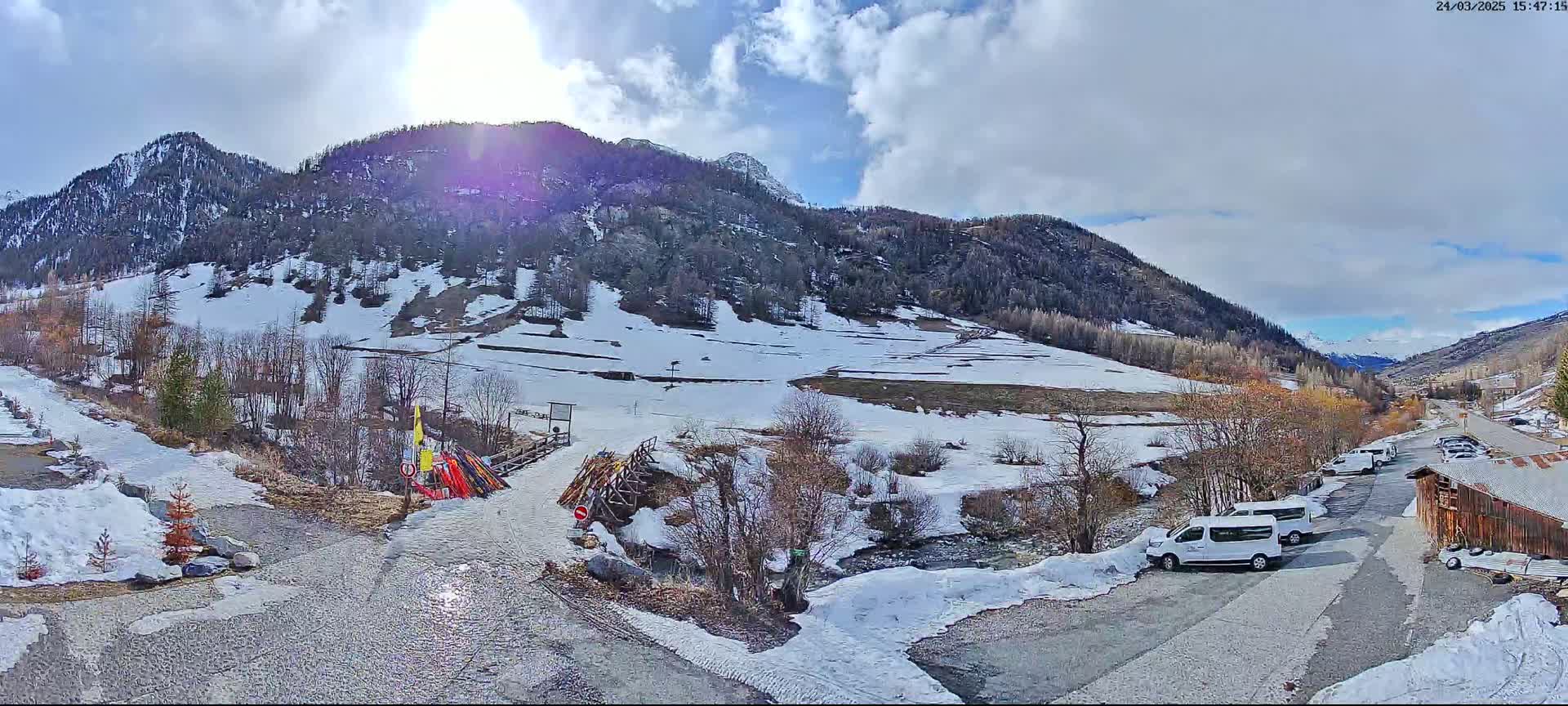 A sunny day in a snow-covered mountain valley shows a small parking area with several white vans near a rustic wooden building, with a partially melted stream flowing through the valley.