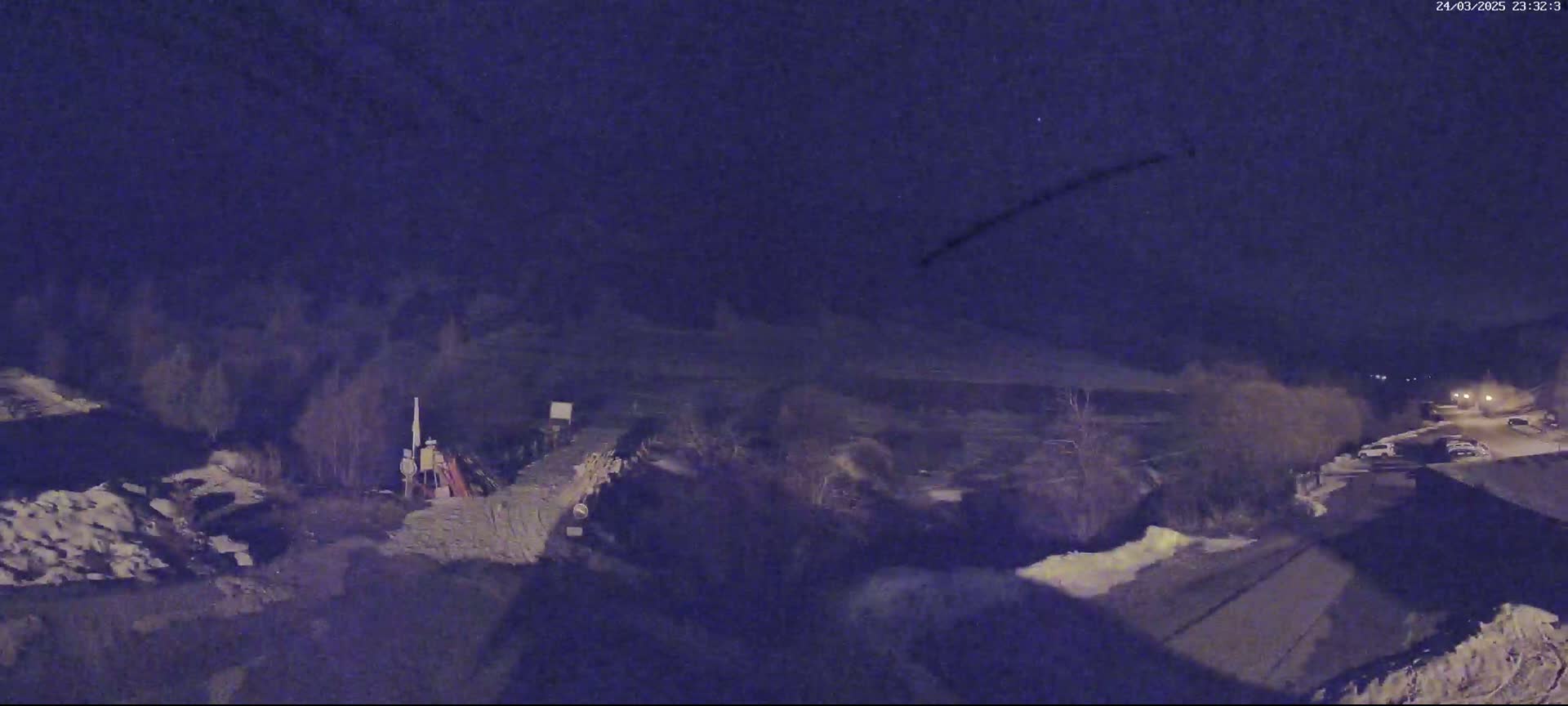 A nighttime, high-angle view shows a dark landscape with patches of snow, some sparse trees, and a road with a few parked cars visible under a dark, mostly clear sky.
