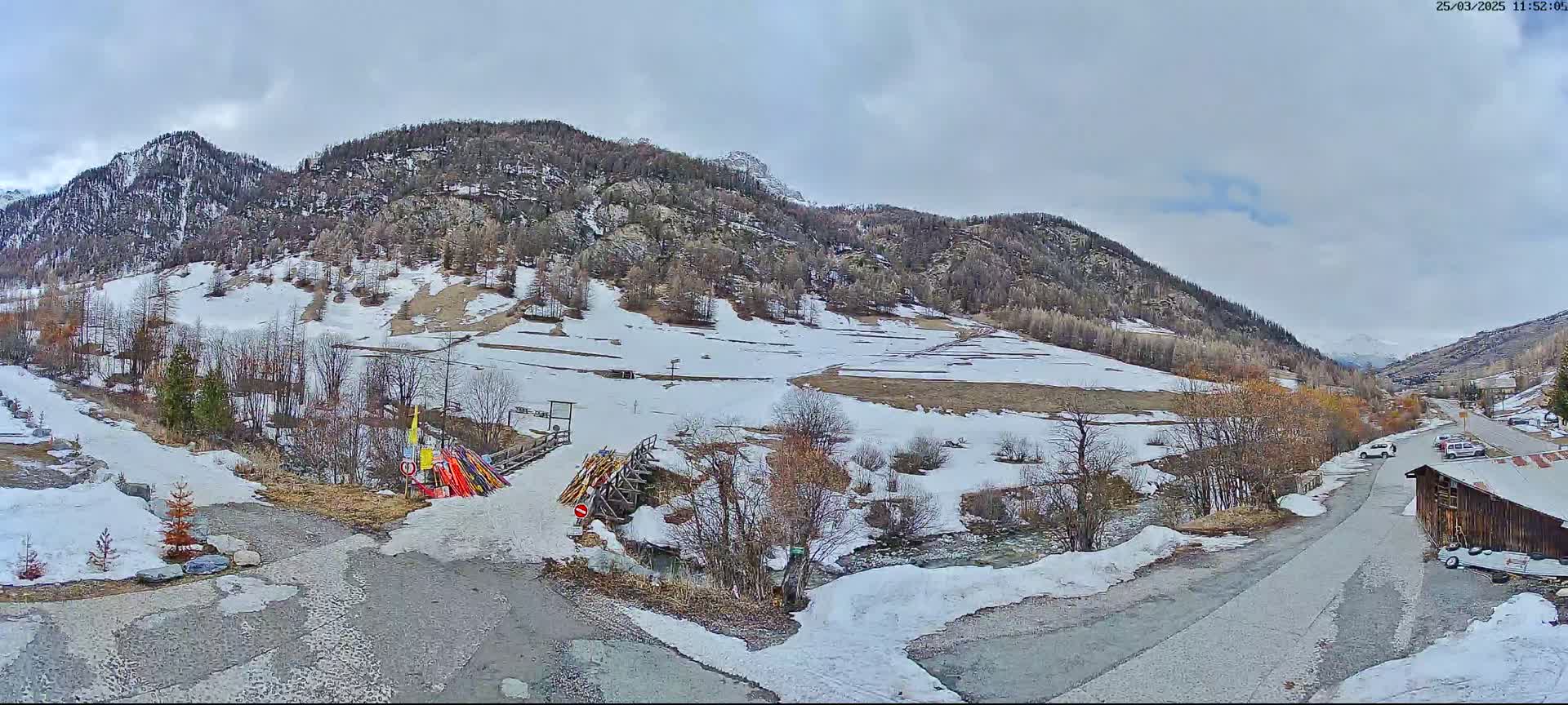 A snow-dusted mountain valley scene under a cloudy sky shows a small stream, a road, and a few parked cars near some buildings.