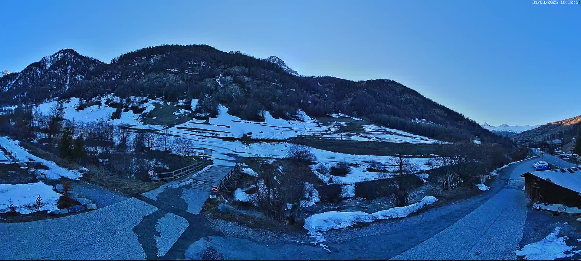 A snow-dusted mountain valley road leads to a small wooden structure, with a wooden bridge crossing a partially frozen stream, under a clear blue sky.